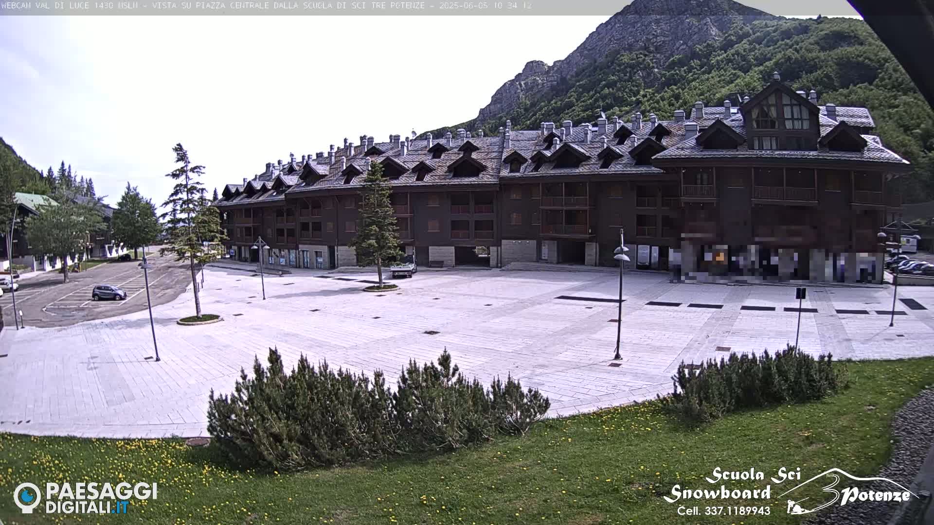 A large, empty paved square sits in front of a multi-story brown building nestled in a mountain valley under a mostly sunny sky.