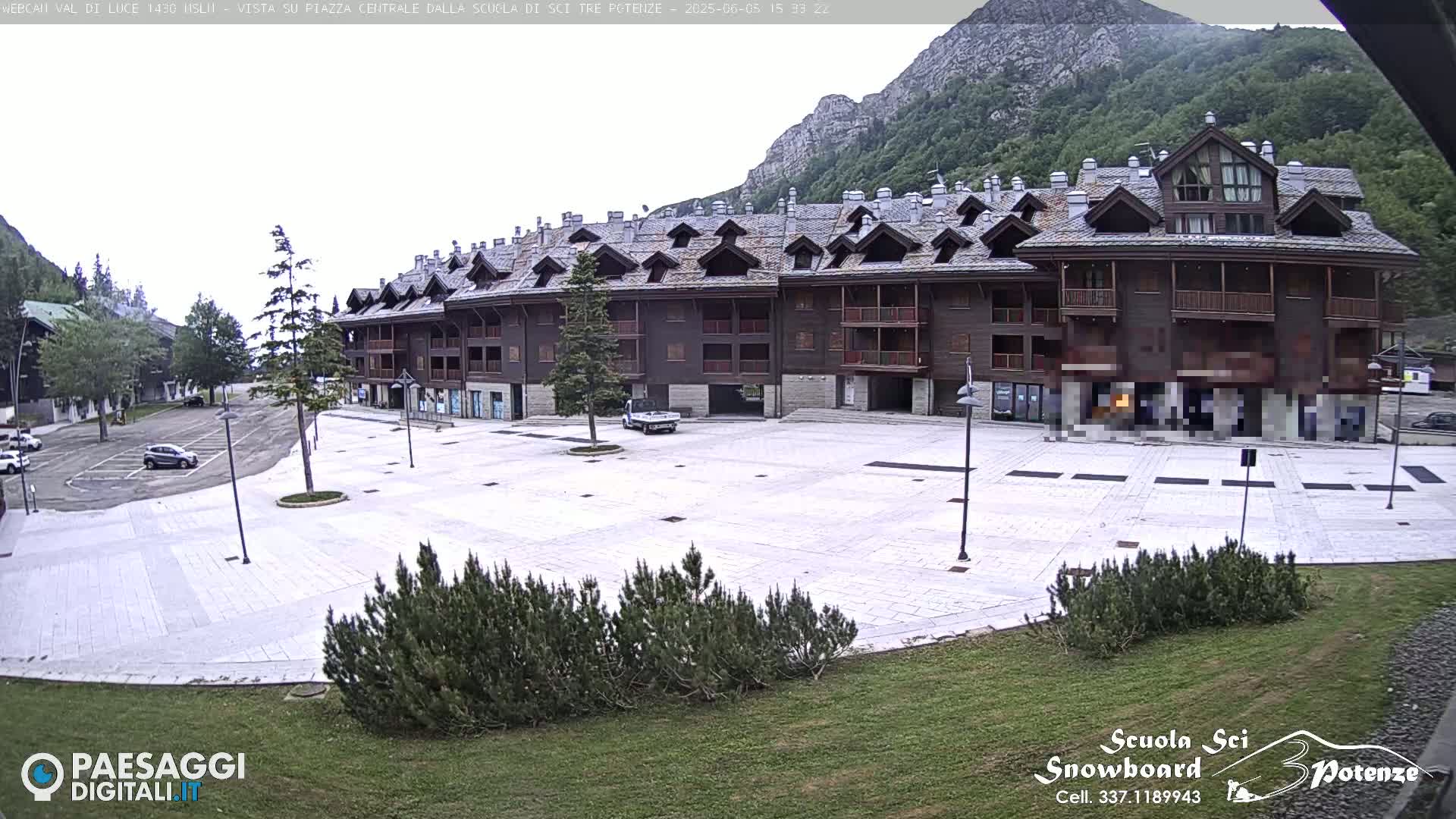 A large, empty plaza paved with light-colored stones sits in front of a multi-story, dark brown building in a mountainous area under an overcast sky.