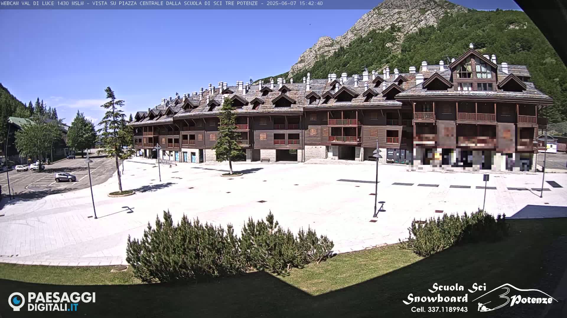 A large, light-colored plaza sits in front of a multi-story brown building, all under a clear, sunny sky, with mountains visible in the background.