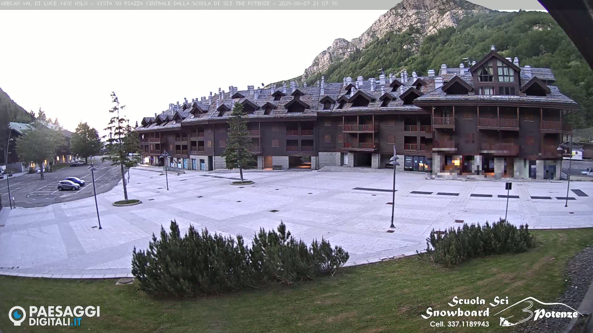 A large, paved plaza sits in front of a multi-story building, with mountains in the background under a partly cloudy sky.