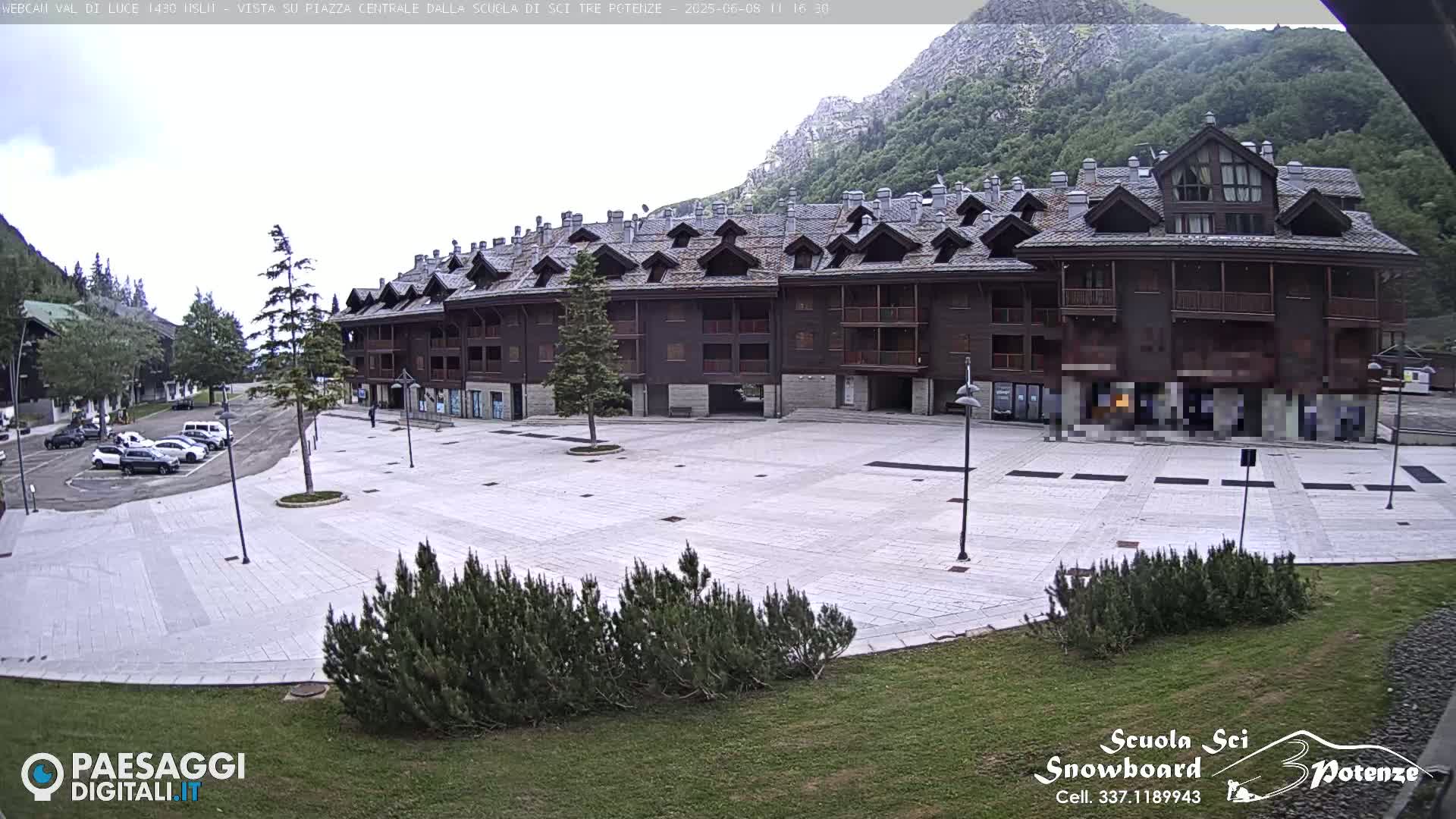 A large, light-colored plaza sits in front of a multi-story brown building against a backdrop of mountains under an overcast sky.