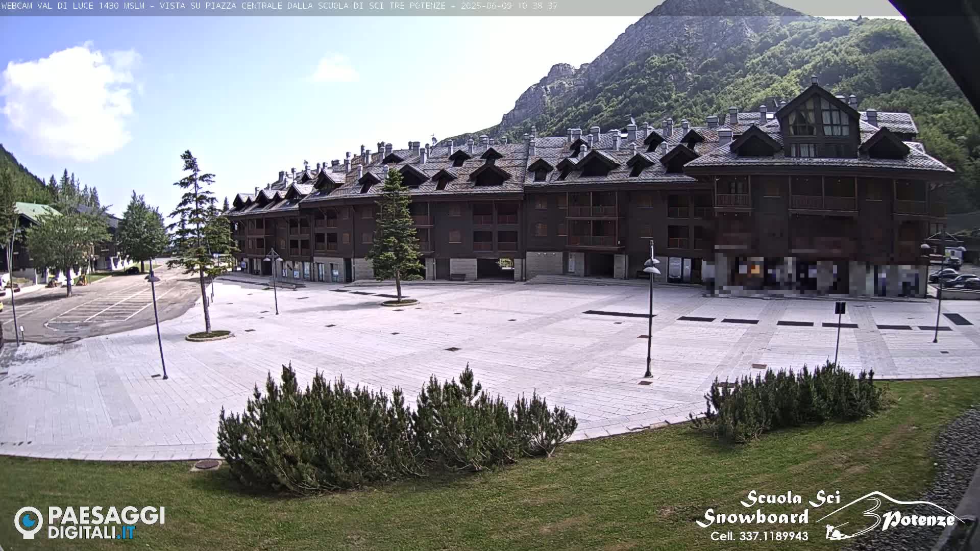 A large, empty plaza paved with light-colored stone is situated in front of a multi-story brown building, with mountains and a clear sky in the background.