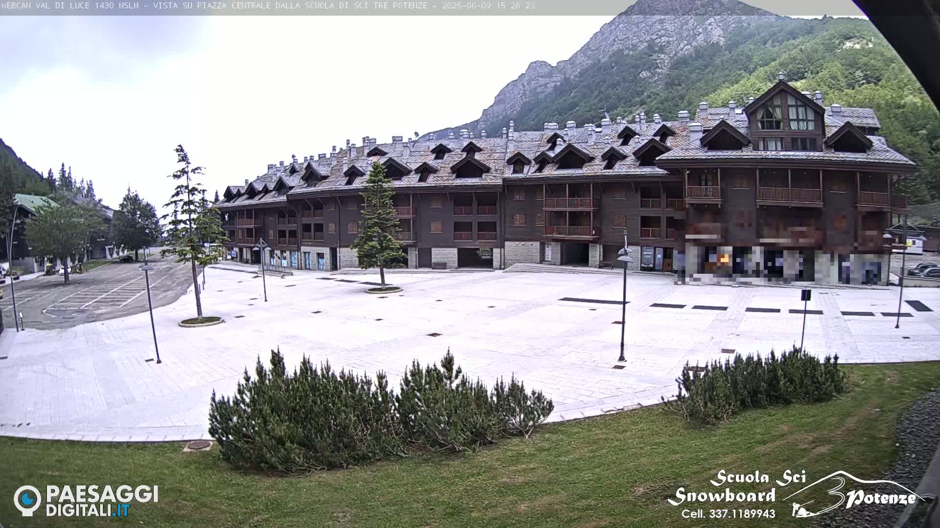 A large, light-colored paved plaza sits in front of a multi-story building with a dark wood exterior and steeply pitched roofs, all set against a backdrop of mountains under an overcast sky.
