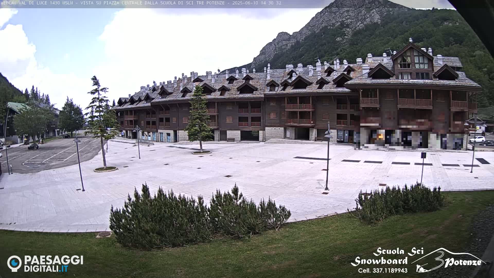 A large, light-colored paved plaza sits in front of a multi-story, dark brown building with many windows and balconies, all under a partly cloudy sky with mountains in the background.
