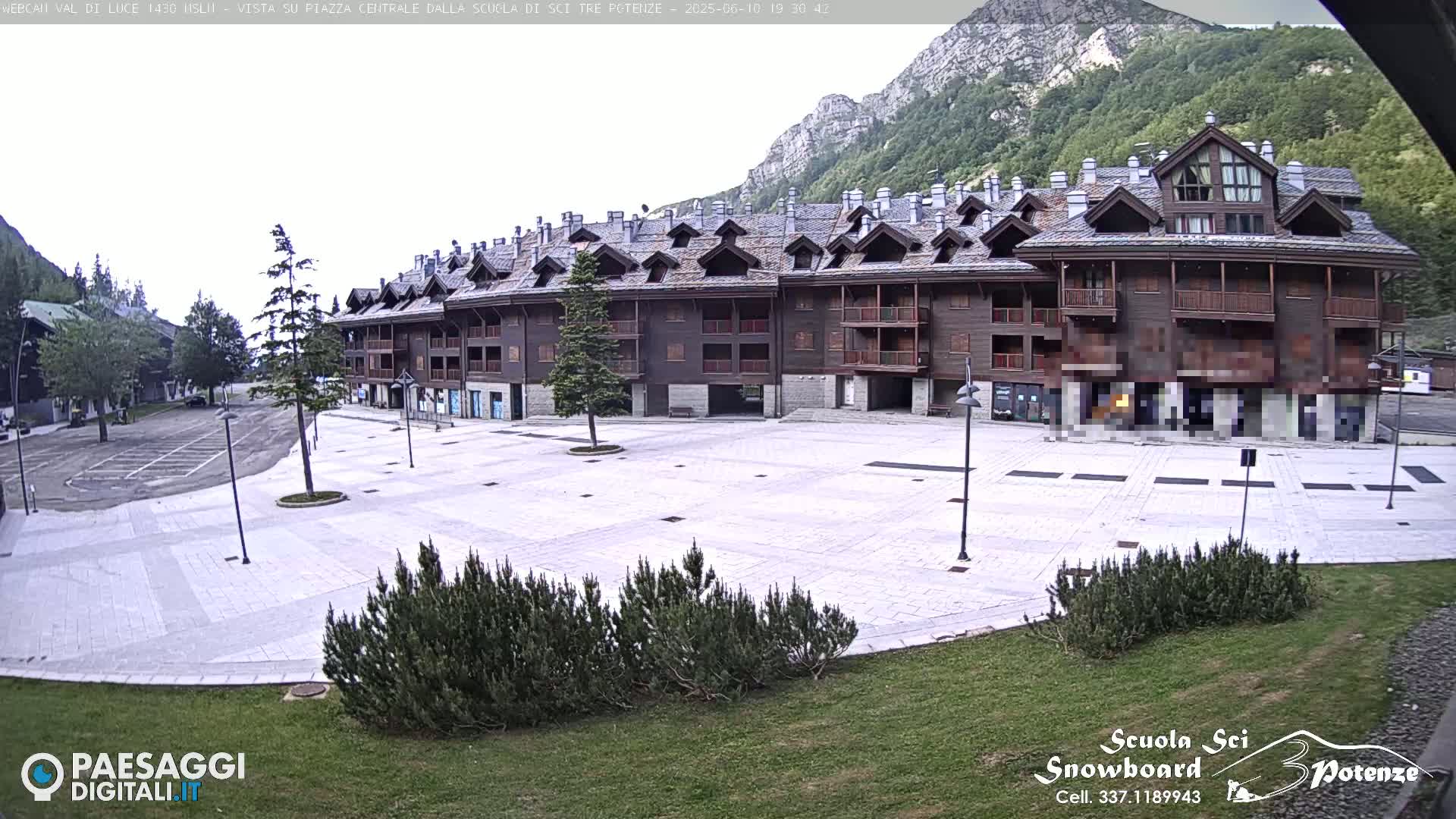 A large, empty plaza paved in light-colored stone is situated in front of a multi-story, dark brown wooden building against a backdrop of mountains under an overcast sky.