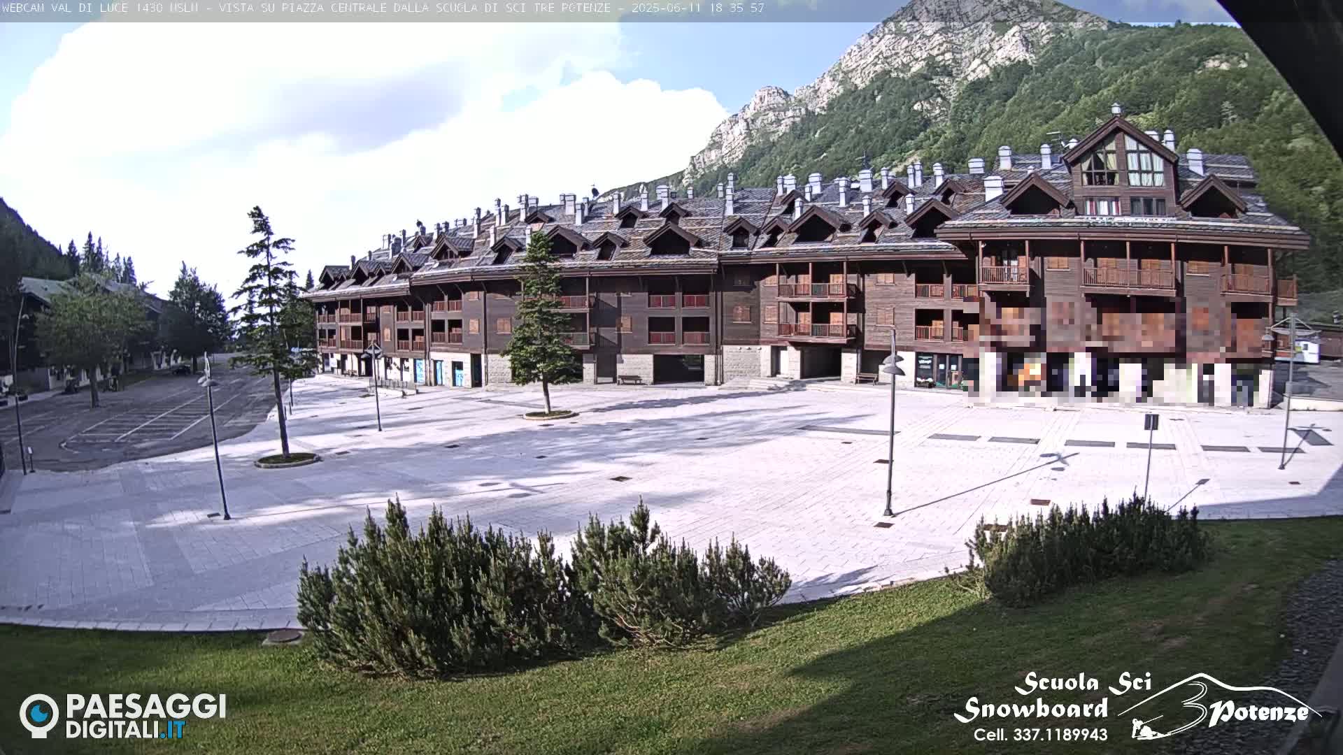 A large, empty plaza paved in light-colored stone is situated in front of a multi-story wood-sided building, with mountains and a mostly clear sky in the background.