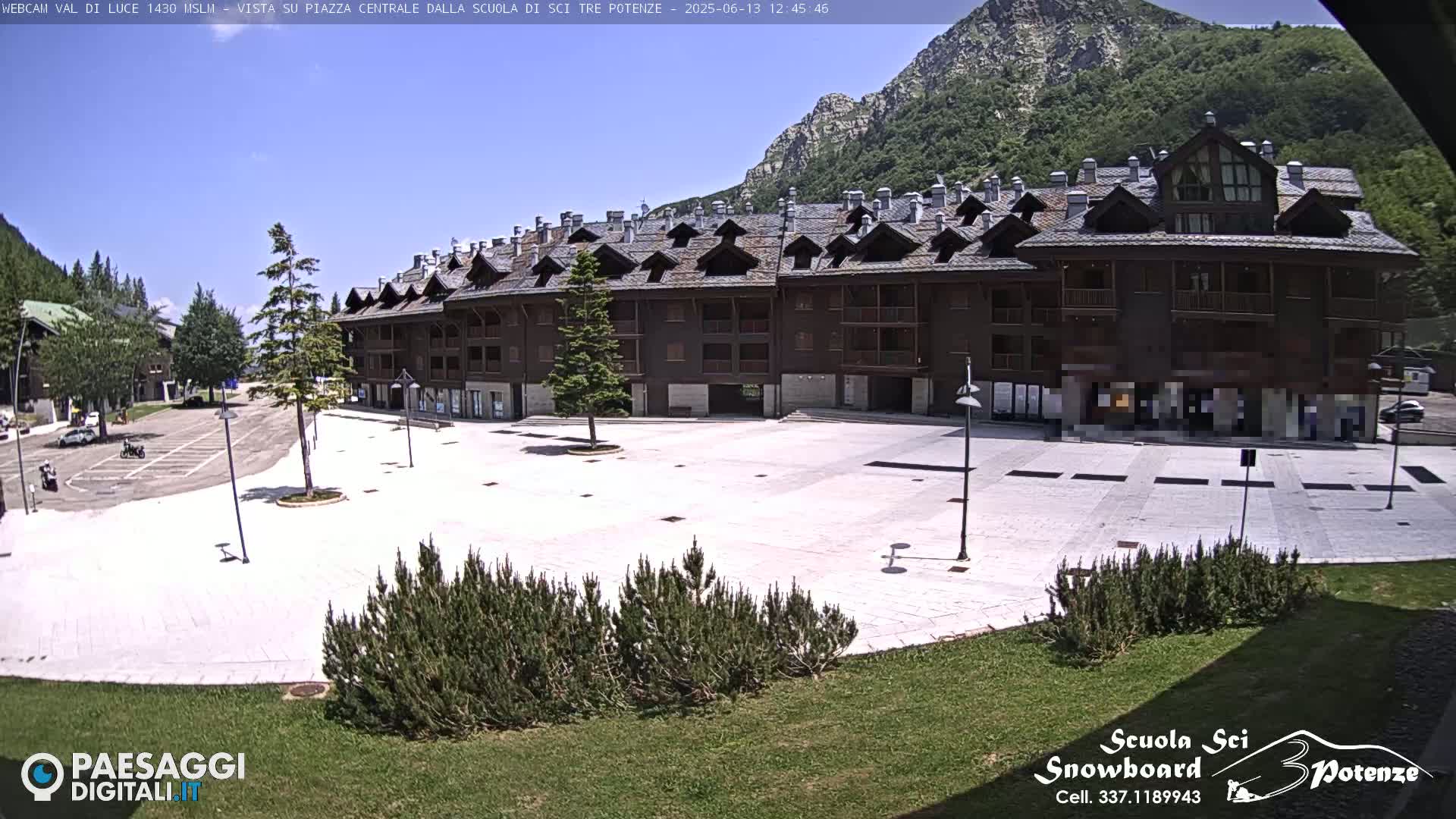 A large, empty plaza paved in light-colored stone sits in front of a multi-story, dark brown building, all under a bright, sunny sky with mountains in the background.