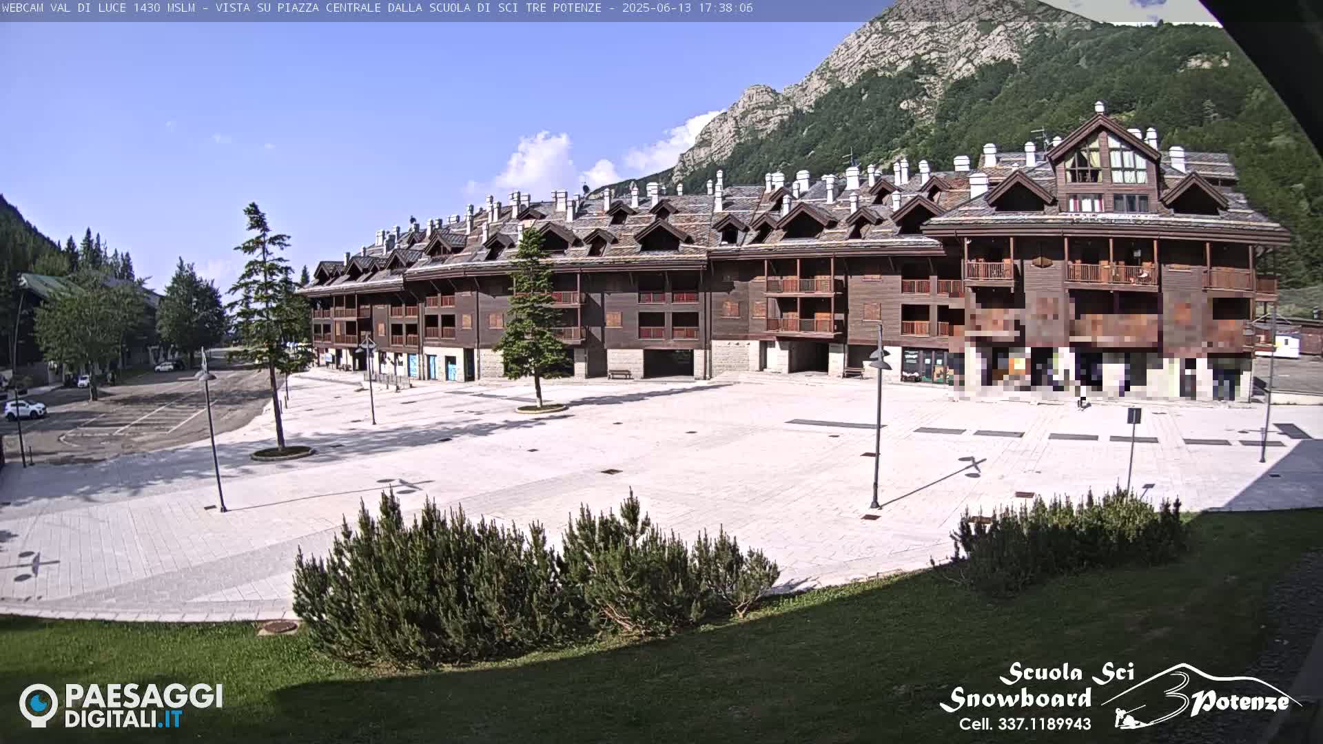 A mostly empty paved plaza sits in front of a large, dark-brown multi-story building, with mountains and a partly cloudy sky in the background.