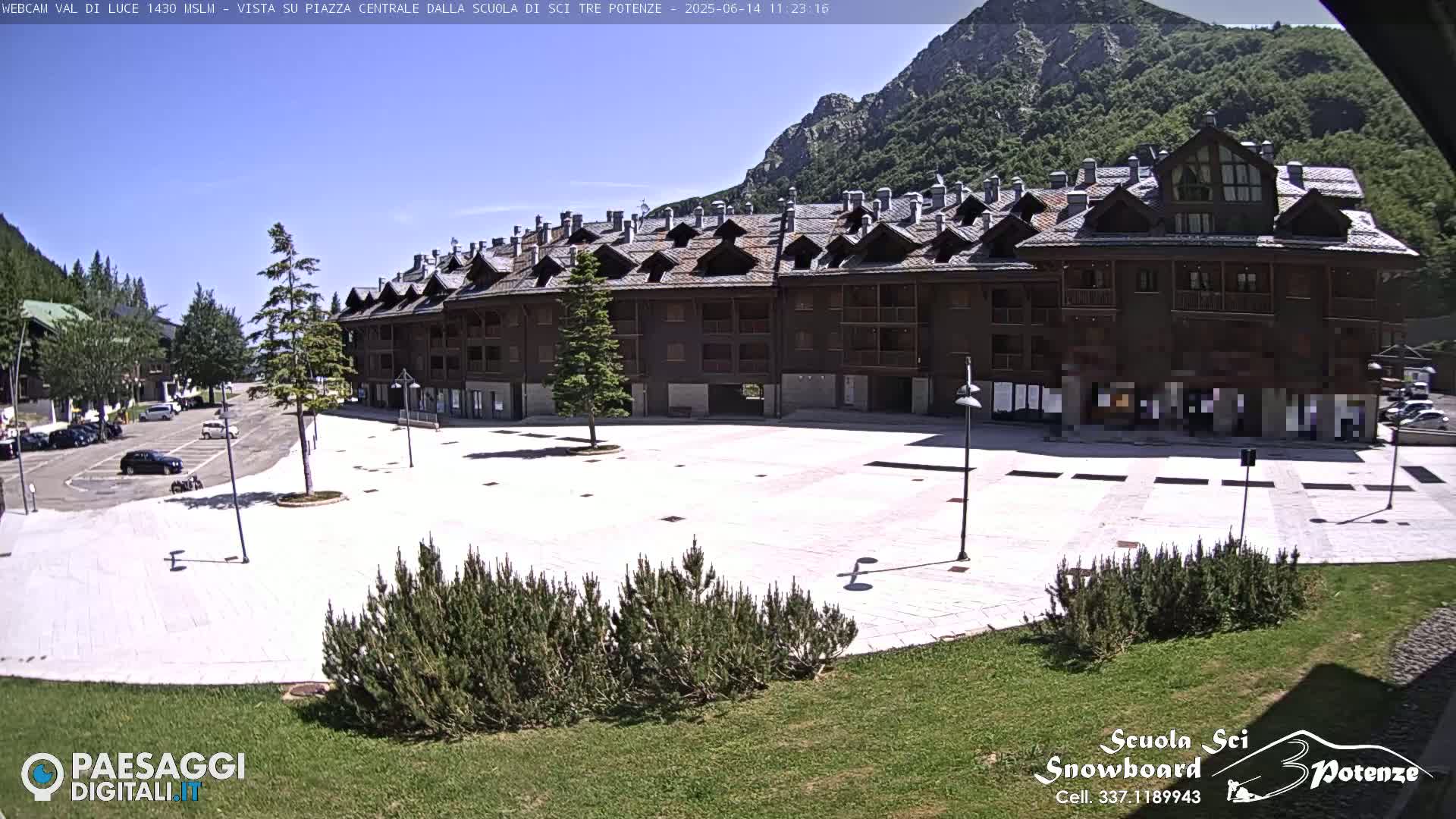 A large, empty plaza with a multi-story building is seen on a sunny day, with mountains in the background.
