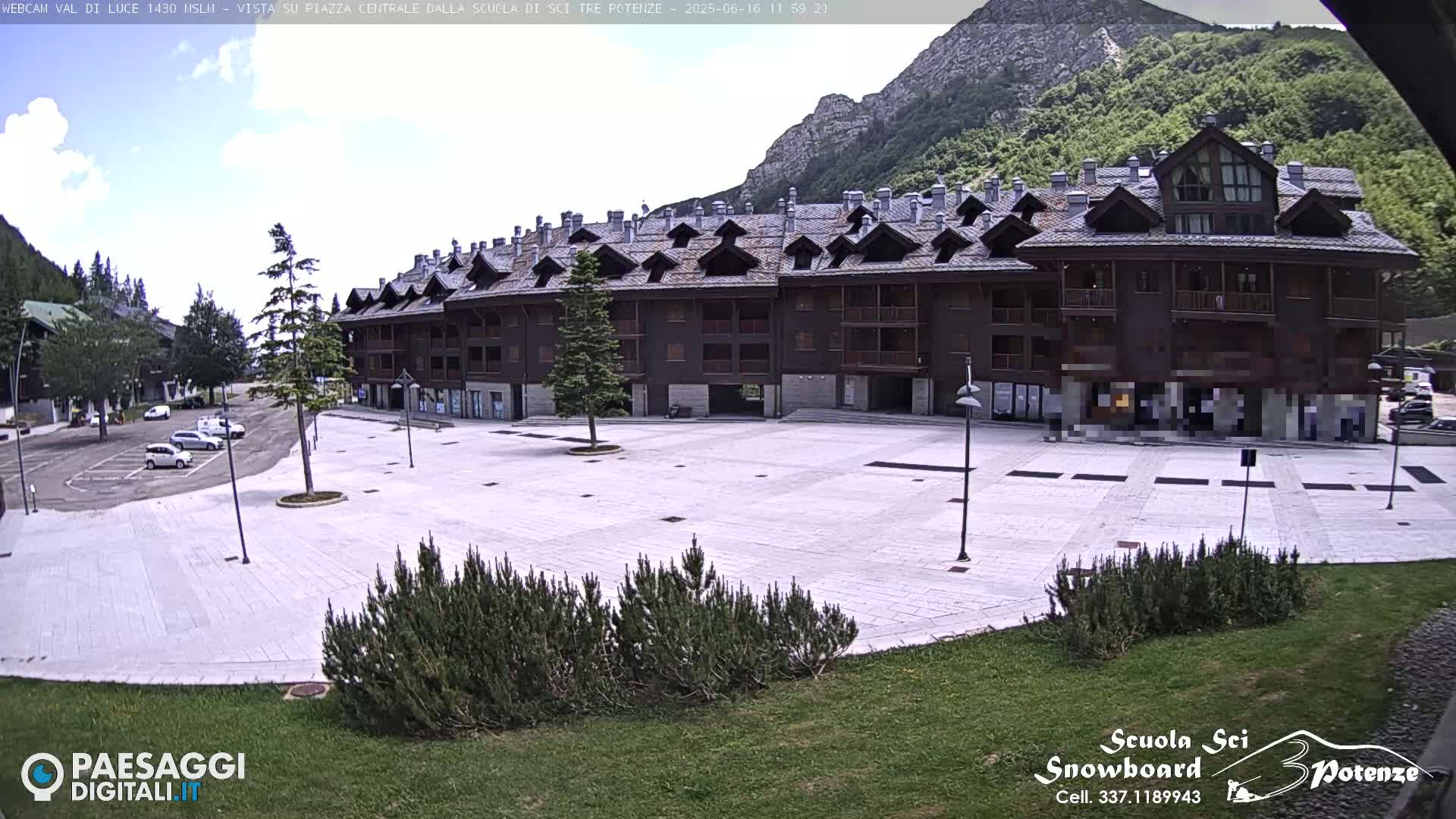 A large, empty plaza paved in light-colored stone sits in front of a multi-story, dark-brown building against a backdrop of mountains under a partly cloudy sky.