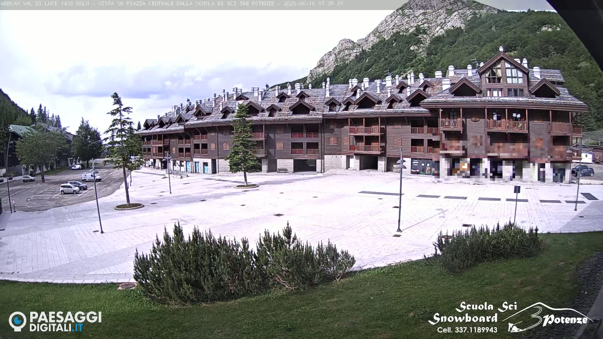 A large, multi-story brown building sits in a town square under a partly cloudy sky, with mountains visible in the background.