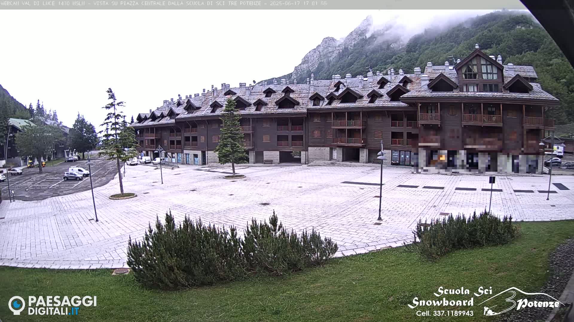 A large, stone plaza sits in front of a multi-story, wood-sided building, with mountains visible in the background under an overcast sky.