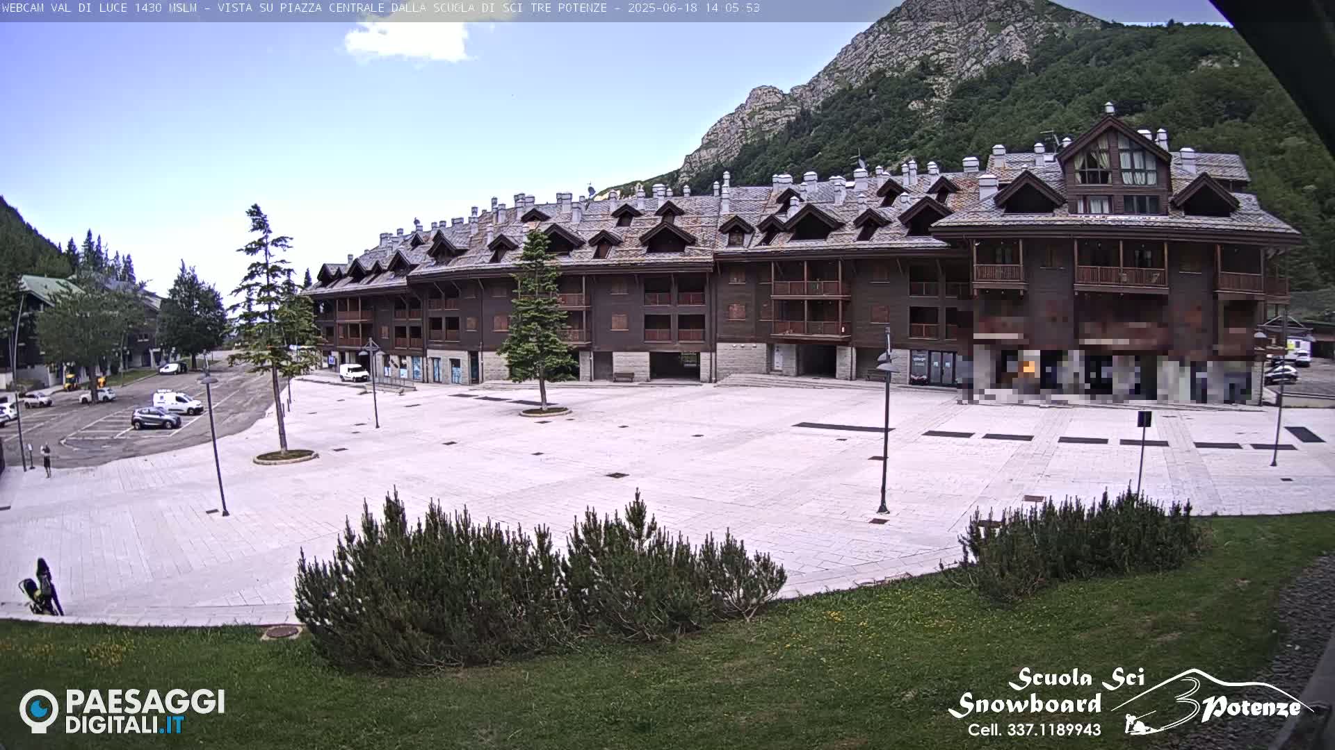 A mostly empty paved plaza sits in front of a large, multi-story brown building, with a mountain and clear sky in the background.