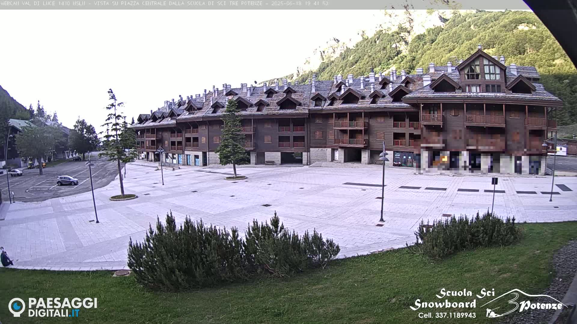 A large, empty plaza paved in light gray stone is situated in front of a multi-story brown building, with mountains and some greenery in the background under a mostly clear sky.