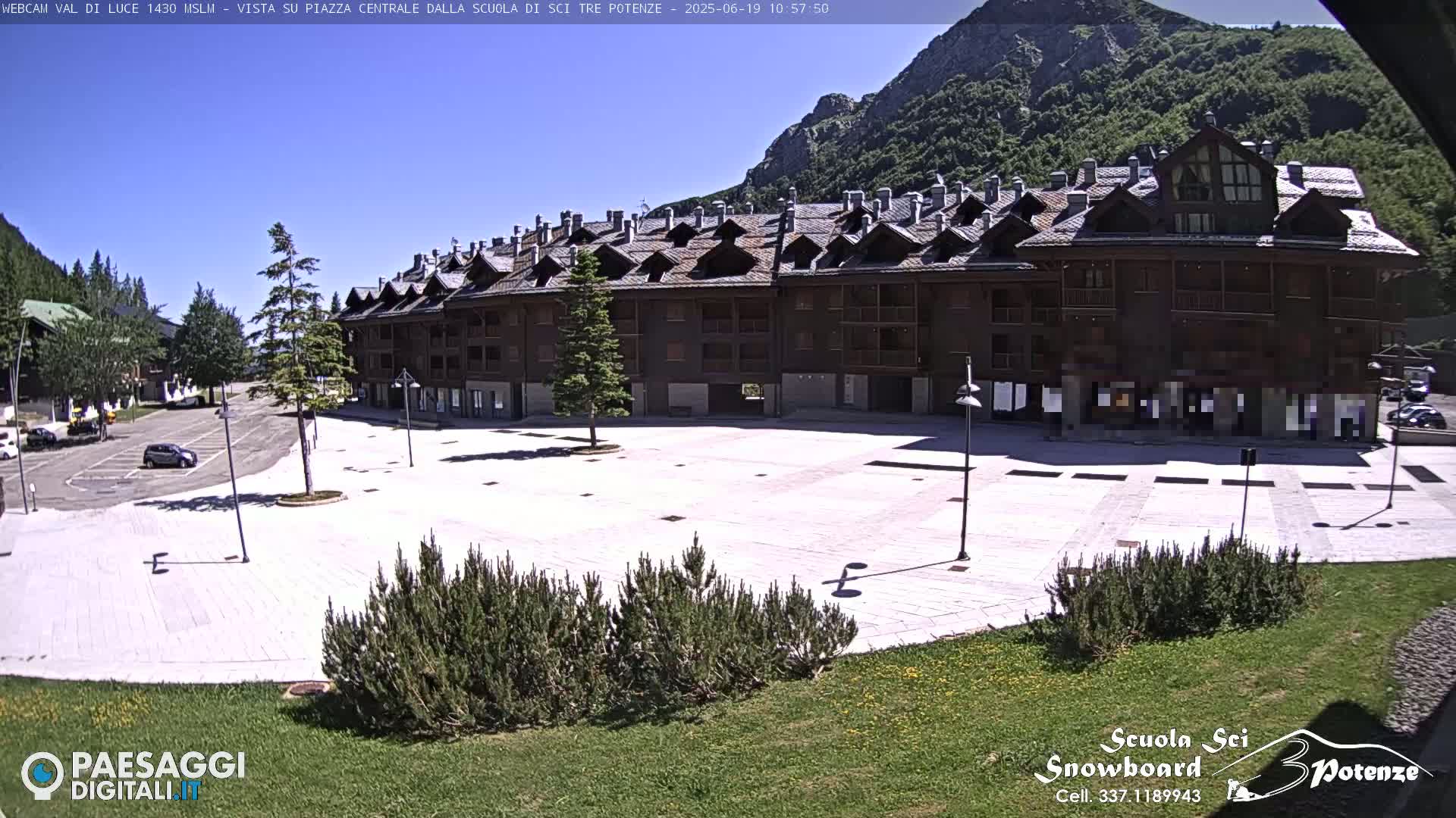 A large, empty plaza paved in light-colored stone sits in front of a long, multi-story brown building, with mountains visible in the background under a clear, sunny sky.
