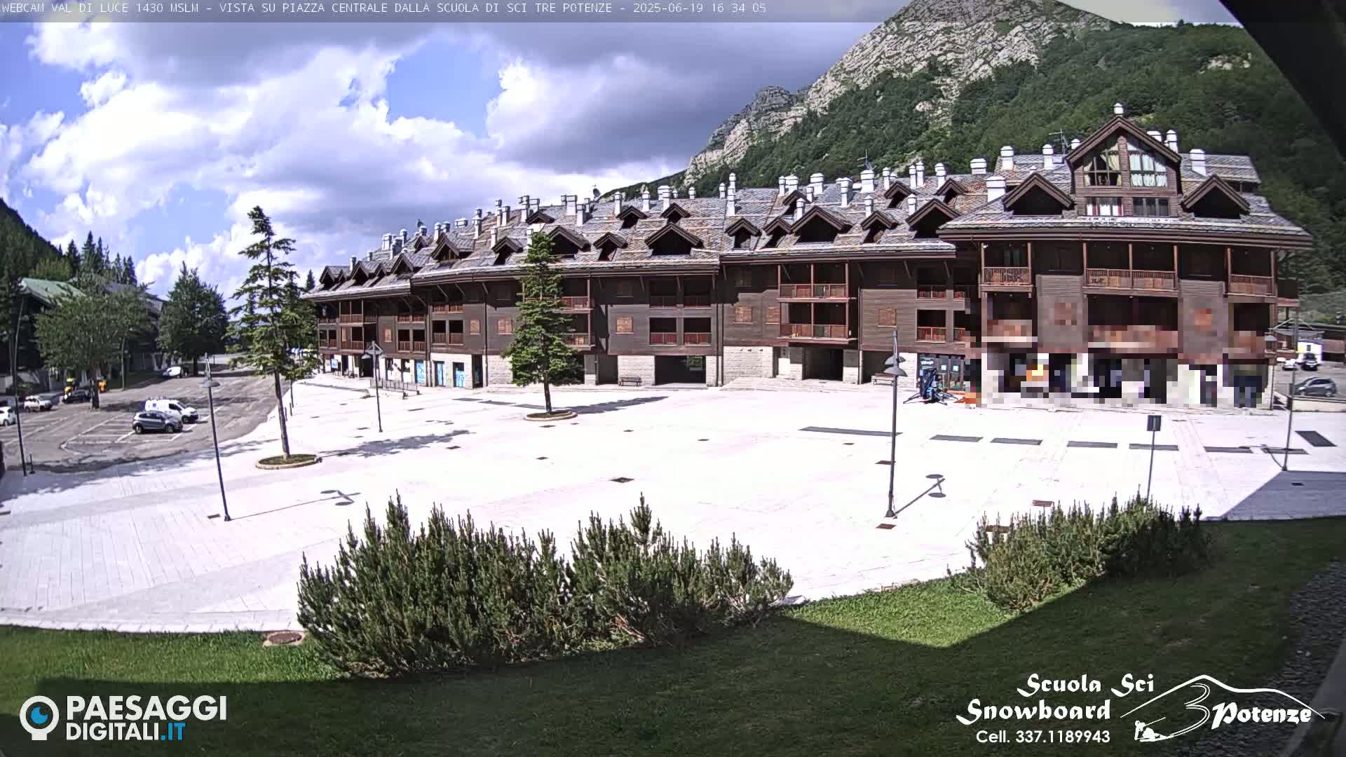 A large, empty plaza sits in front of a multi-story wood-sided building on a partly sunny day with mountains in the background.
