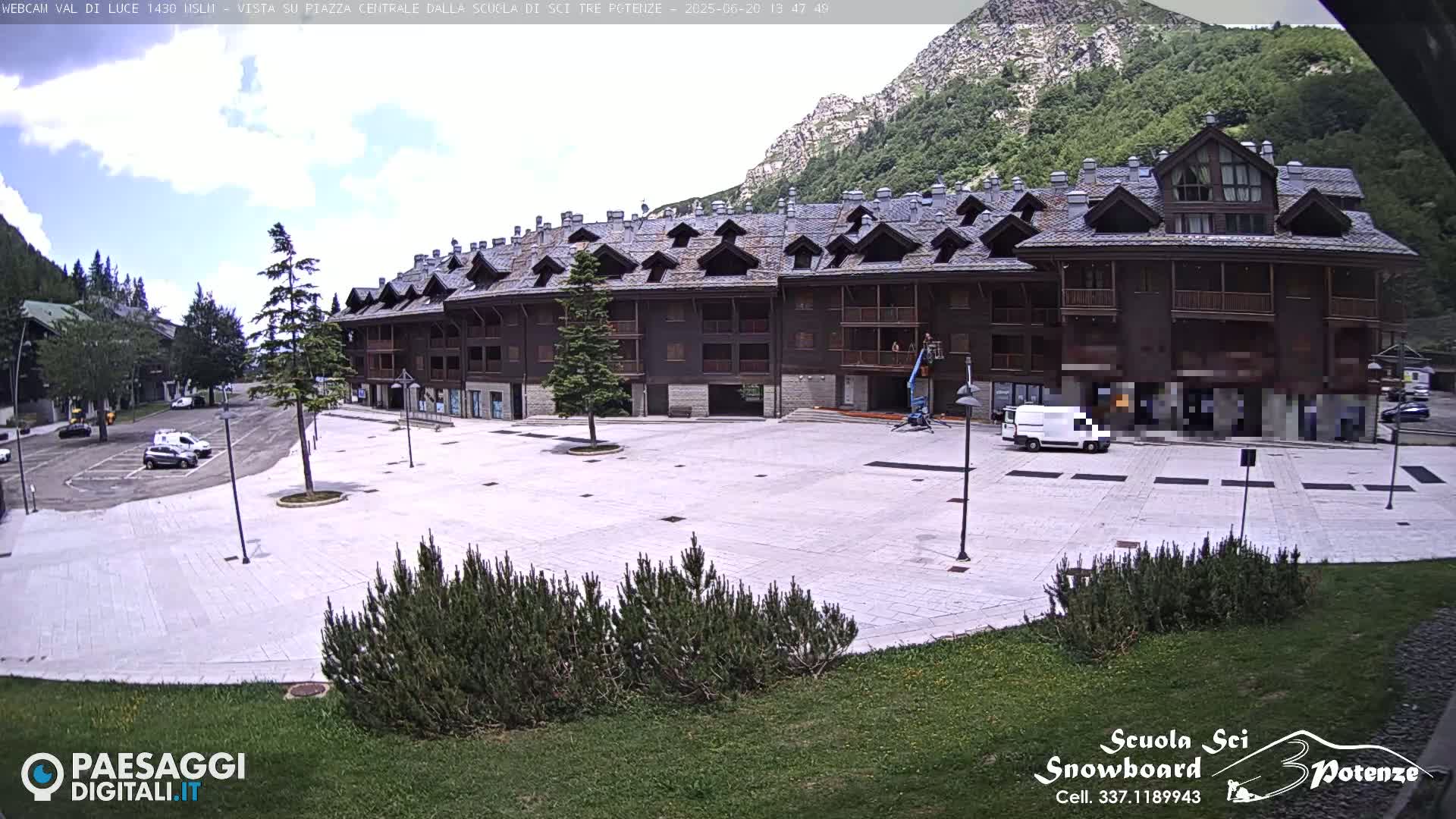 A large, empty plaza paved with light-colored stone is in front of a multi-story brown building, all situated against a mountainous backdrop under a partly cloudy sky.