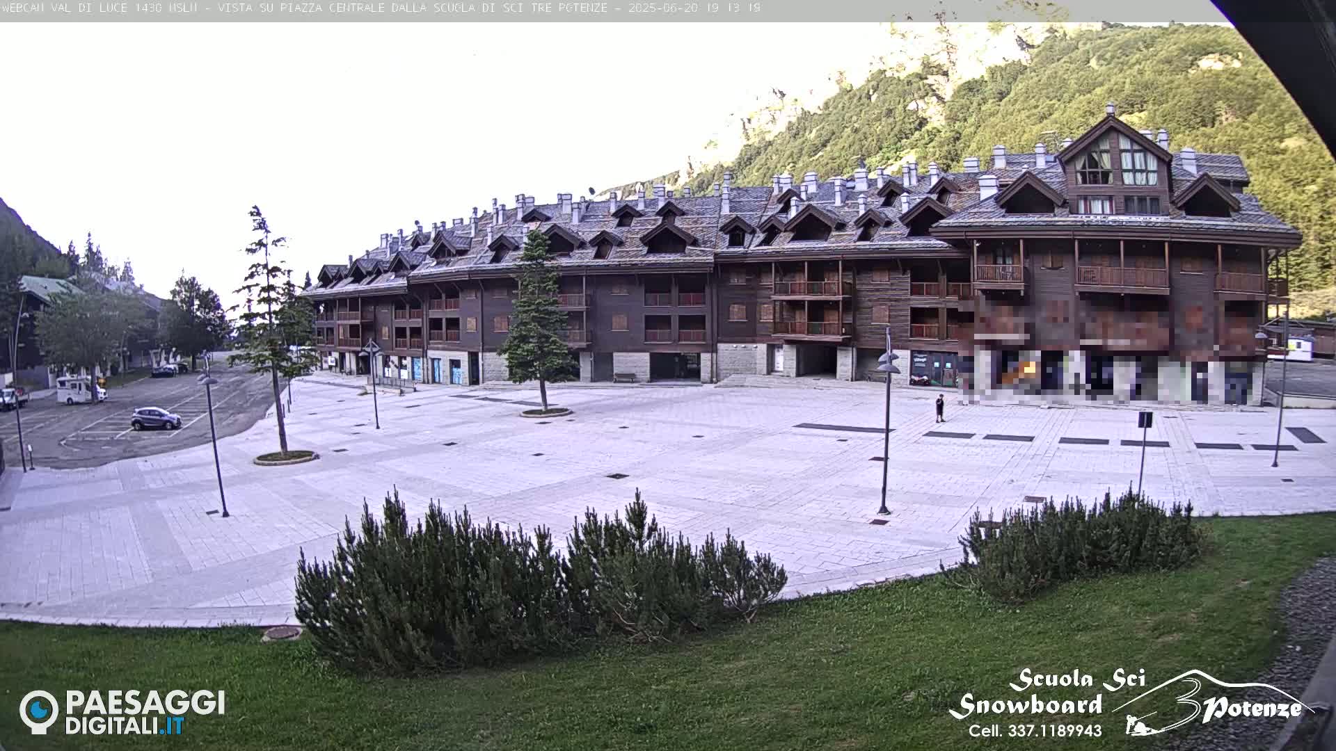 A large, empty plaza is situated in front of a multi-story brown building, with a mountain and forest in the background under a mostly cloudy sky.