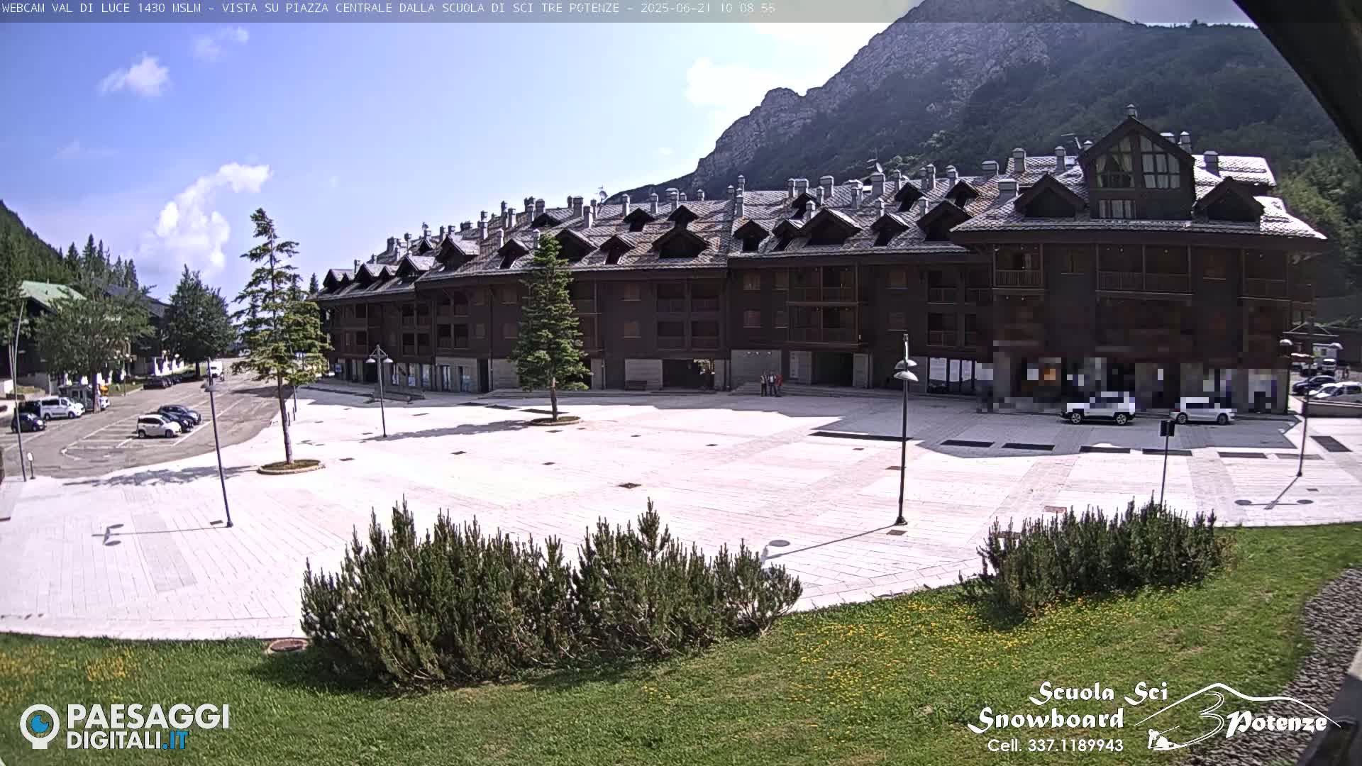 A large, light-colored plaza in front of a multi-story wood-sided building is visible under a partly cloudy sky, with mountains in the background and cars parked nearby.