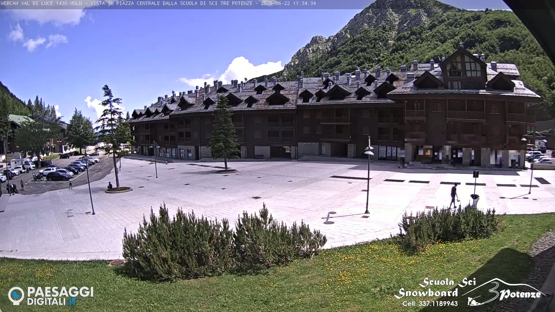 A mostly empty paved plaza is situated in front of a large, multi-story building, with mountains and a partly cloudy sky in the background.