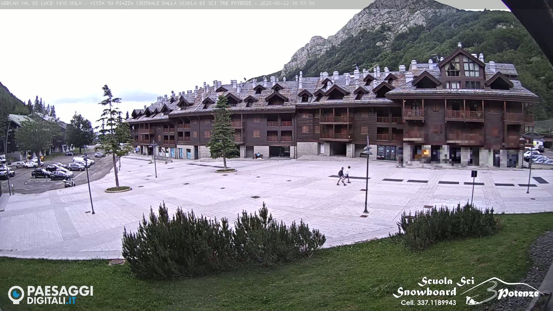 A large, light-grey paved plaza sits in front of a multi-story brown building, with mountains in the background under an overcast sky; cars are parked to the left.