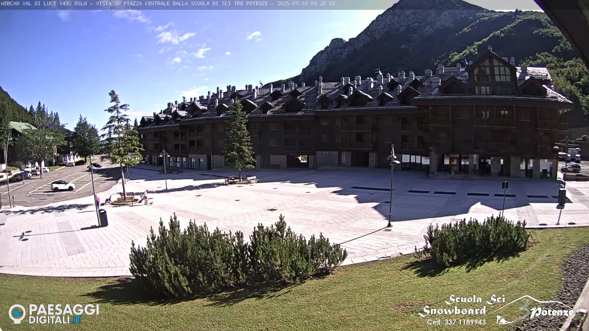 A paved plaza sits in front of a multi-story building in a mountainous region under a mostly sunny sky.