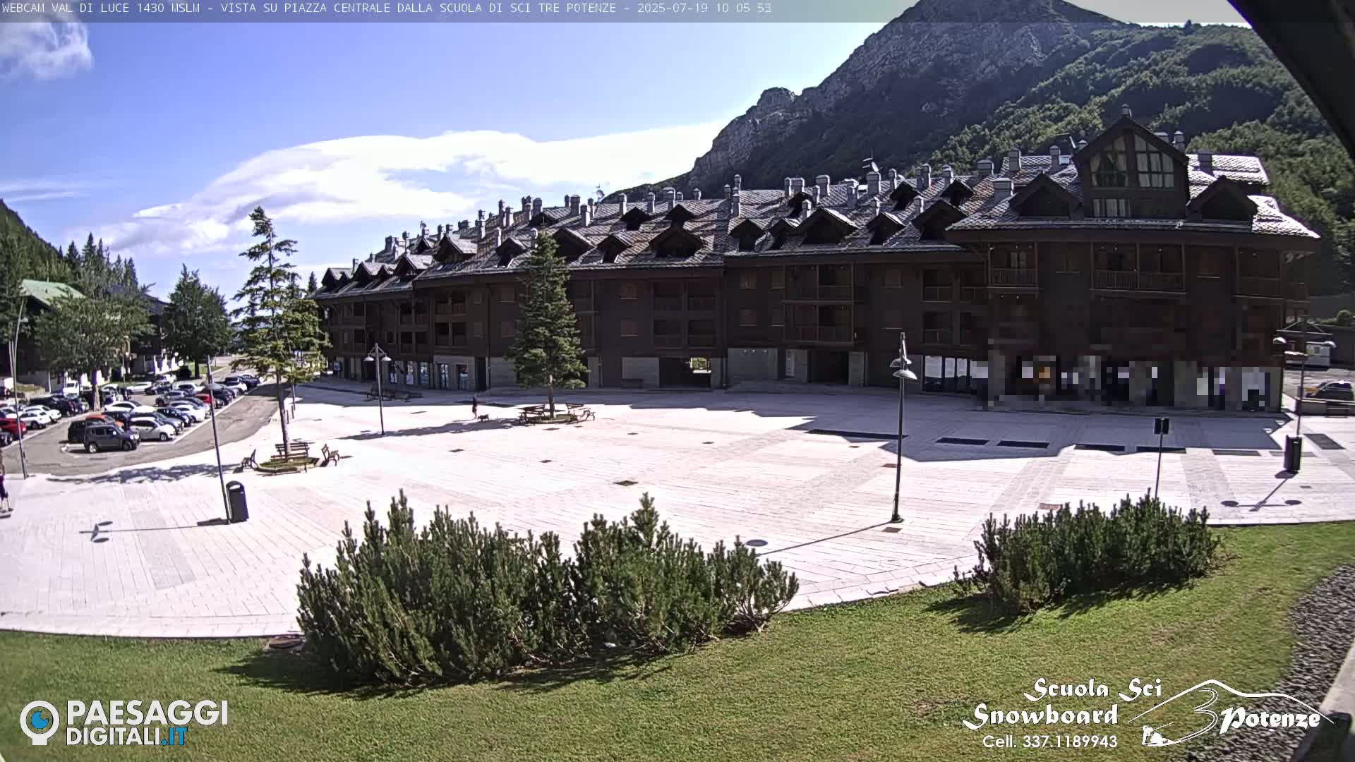 A large, paved plaza sits in front of a multi-story building in a mountainous area under a mostly sunny sky.
