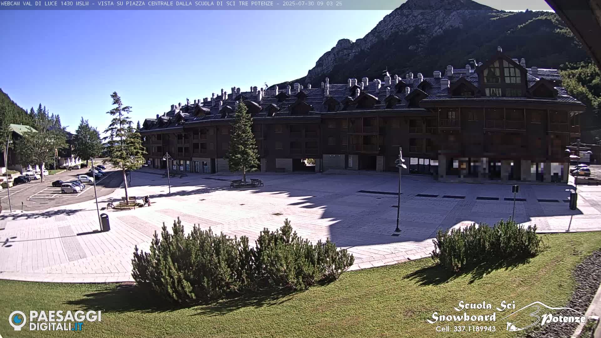 A sunny day shows a paved town square with a large, dark-brown multi-story building behind it, a few cars parked nearby, and some low landscaping.