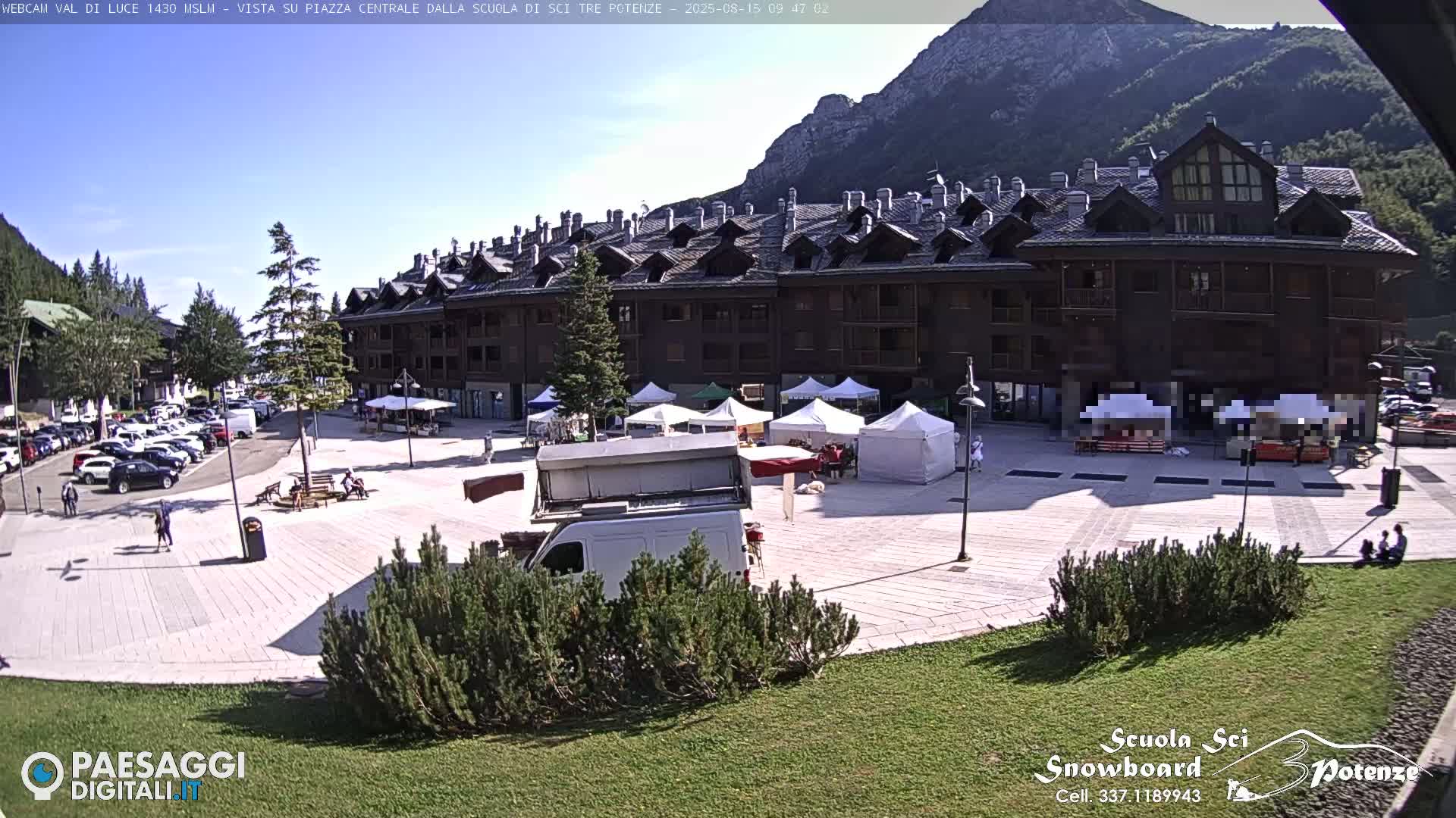 A sunny day shows a town square with several market stalls set up, a building in the background, and a parking lot filled with cars.