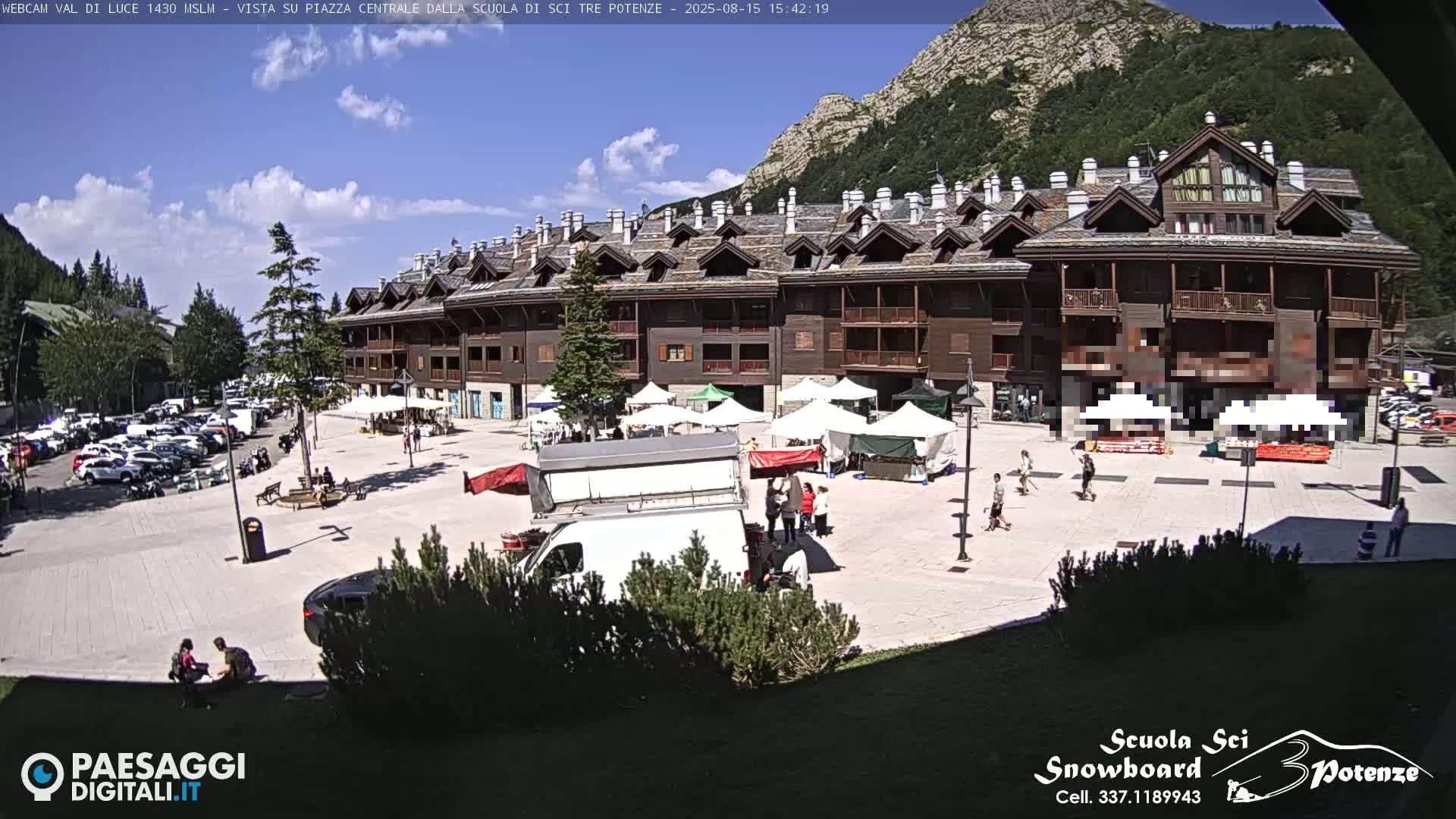 A sunny day shows a town square with several market stalls set up, a building in the background, and a parking lot filled with cars.