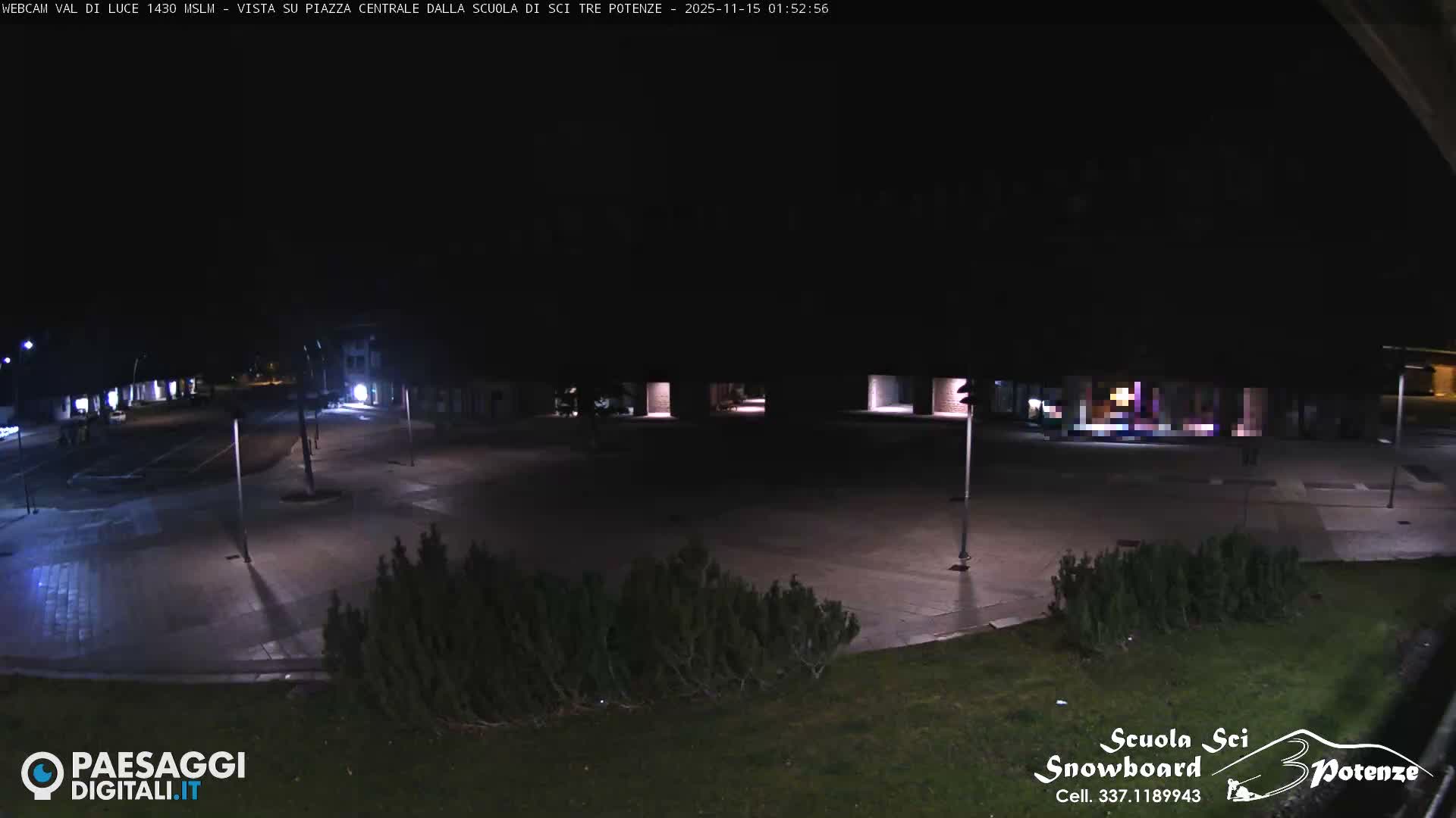 The image depicts a quiet, well-lit town square at night under clear skies, with buildings and some bare trees surrounding a paved area.