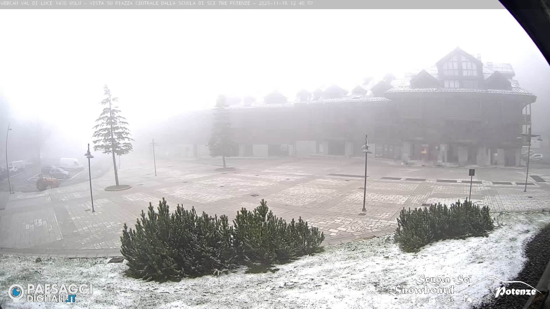 The image depicts a quiet, well-lit town square at night under clear skies, with buildings and some bare trees surrounding a paved area.