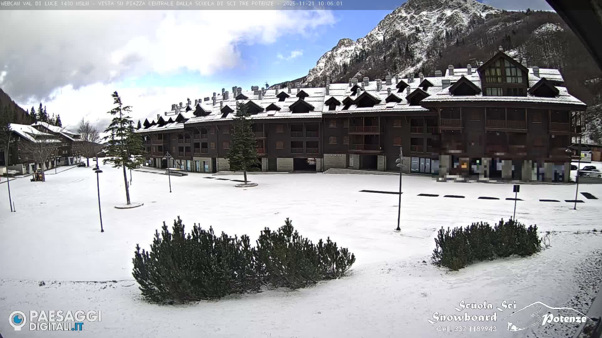 A snow-covered alpine village with dark wooden buildings and evergreen bushes is seen under a partly cloudy winter sky, with snow-capped mountains towering in the background.