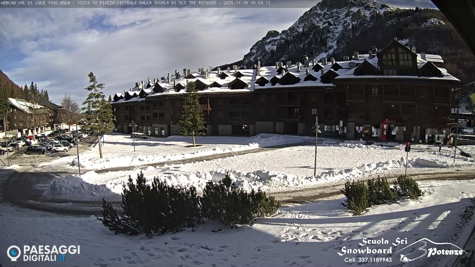 A snowy winter scene depicts a village with dark, multi-story buildings, a parking lot filled with cars, and snow-covered mountains in the background, all beneath a partly cloudy sky.