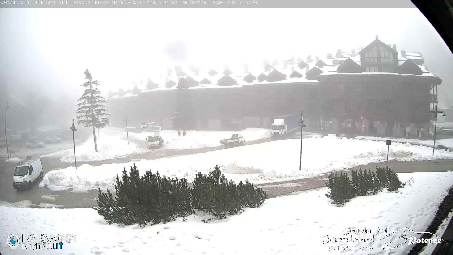 A snow-covered alpine village with a large lodge and several vehicles on partially cleared roads is enveloped in thick fog, severely limiting visibility.