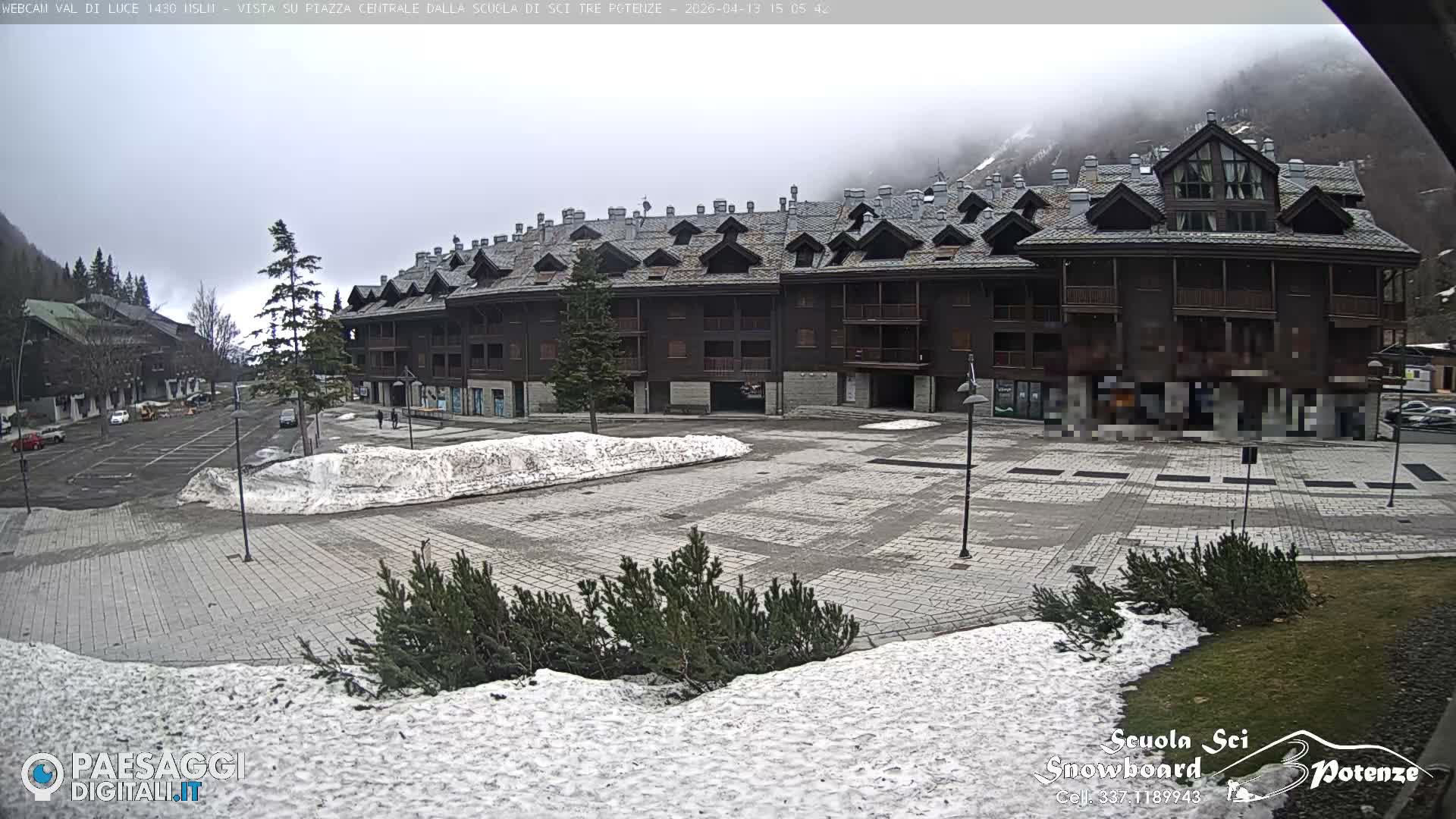 A snow-covered alpine village with a large lodge and several vehicles on partially cleared roads is enveloped in thick fog, severely limiting visibility.