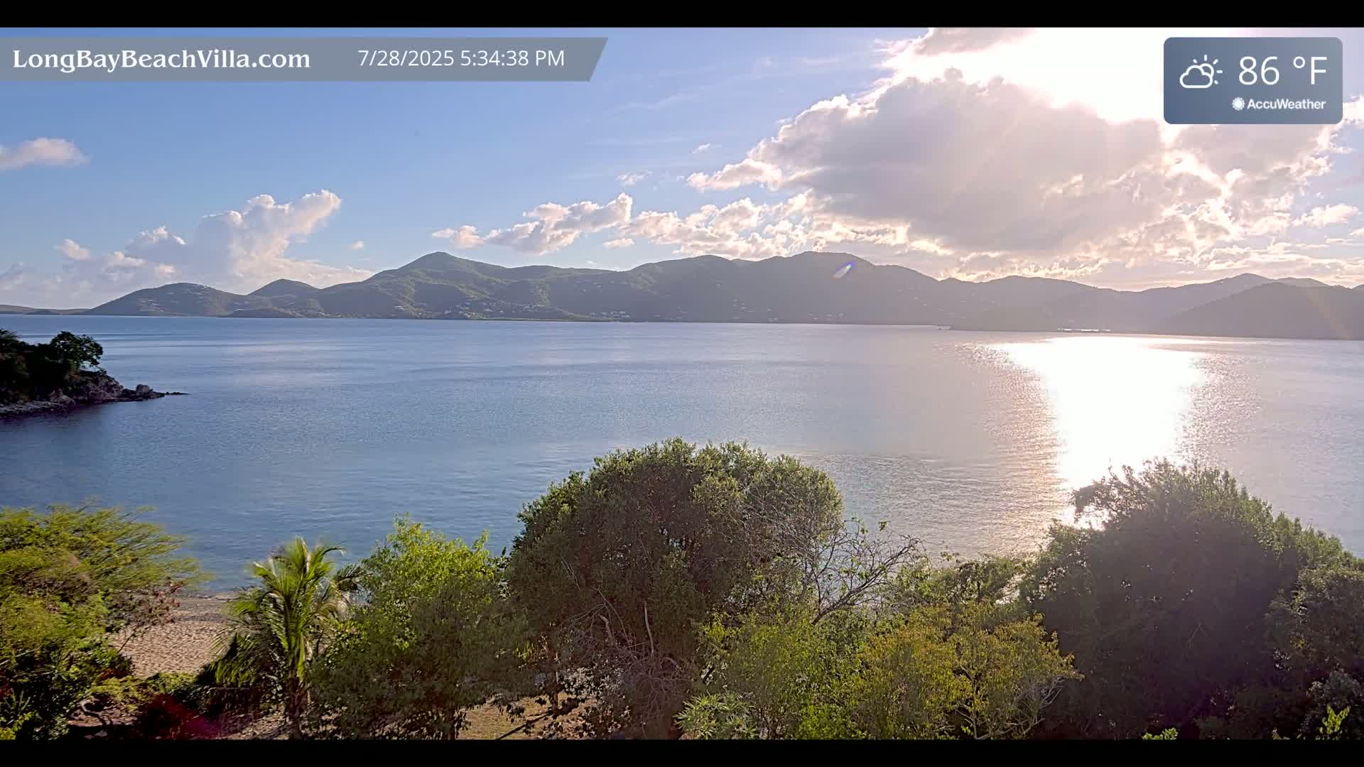 A calm bay with mountains in the background under a partly cloudy sky, with the sun reflecting on the water's surface; the temperature is 86°F.