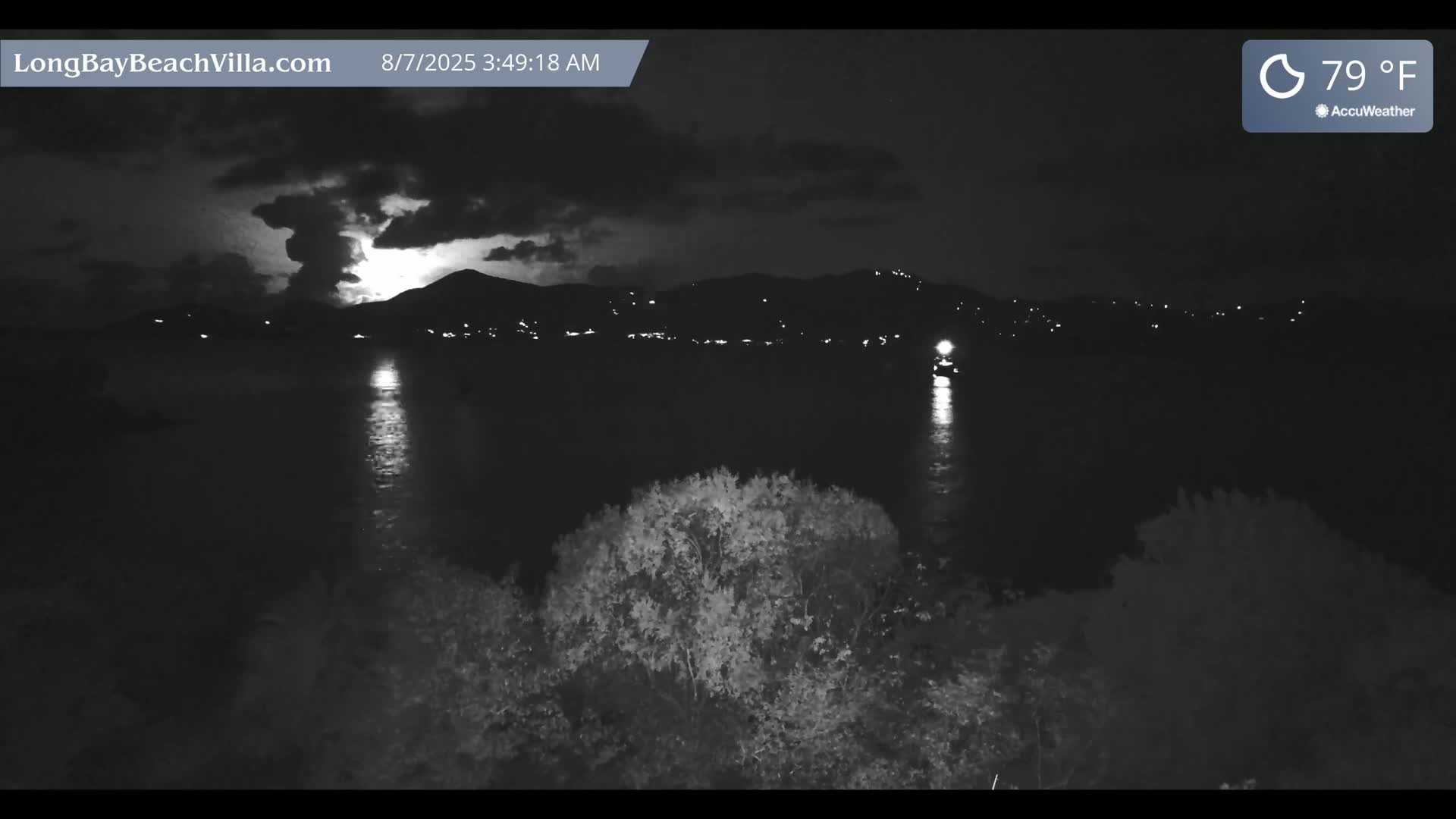 A nighttime view of a calm body of water reflecting lights from a distant shoreline, with a thunderstorm in progress over the mountains in the background.