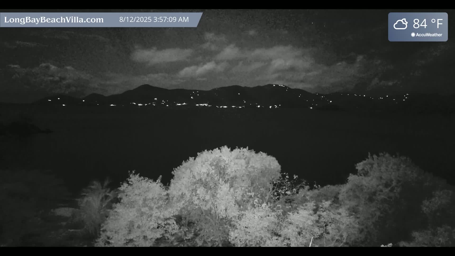 A nighttime view shows a partly cloudy sky, 84 degrees, with distant lights on a mountain range behind a foreground of trees.