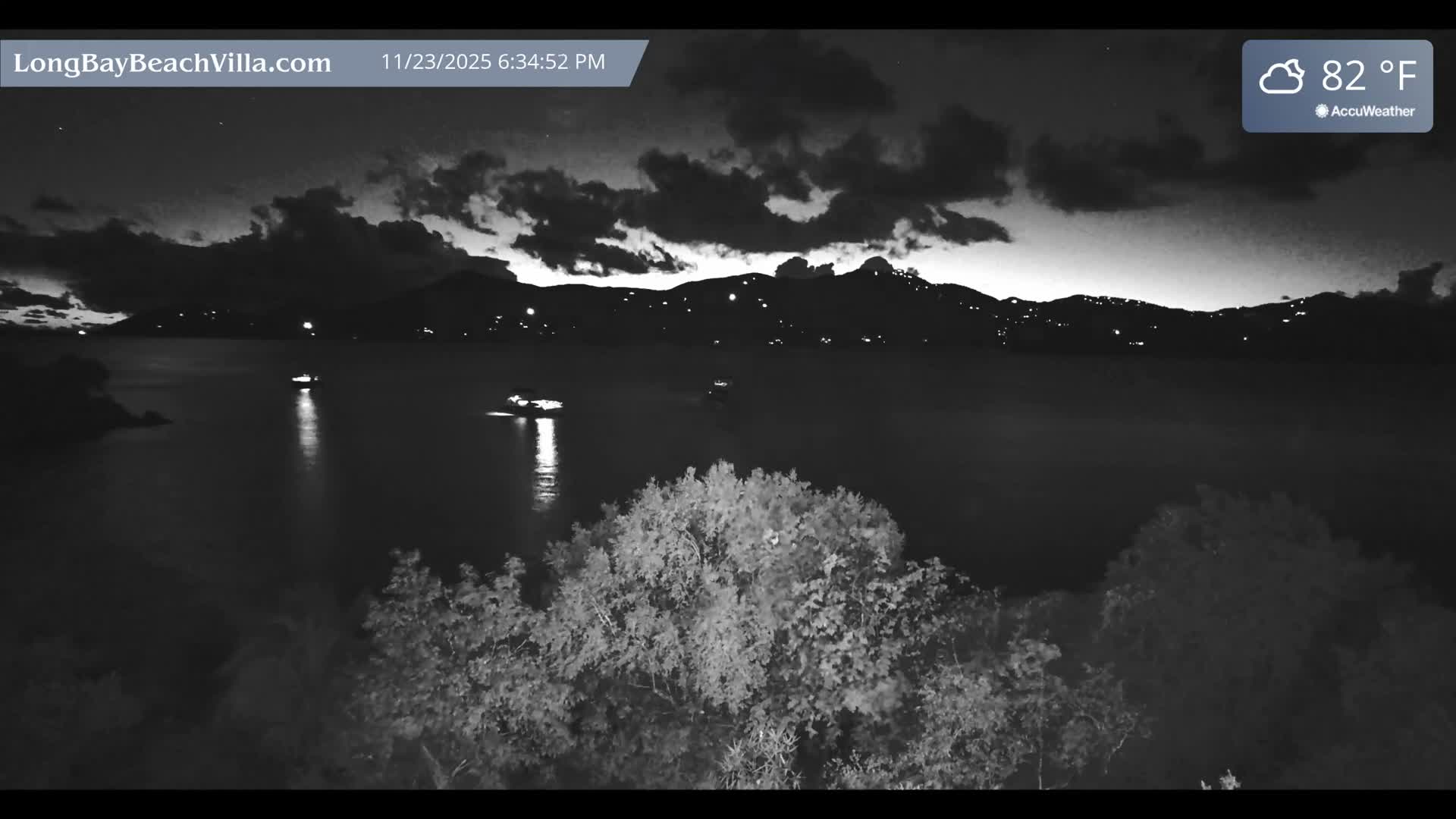The image captures a cloudy night over a calm bay, with several illuminated boats creating reflections on the water, distant mountainous landmasses sparkling with lights, and dense foliage in the foreground, all under an 82°F temperature.