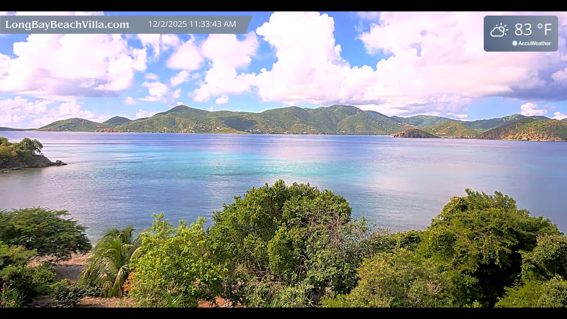 The image displays a picturesque tropical bay featuring calm, clear turquoise and blue waters, framed by lush green hills with scattered buildings under a partly cloudy sky with a temperature of 83°F, and dense green foliage in the foreground.