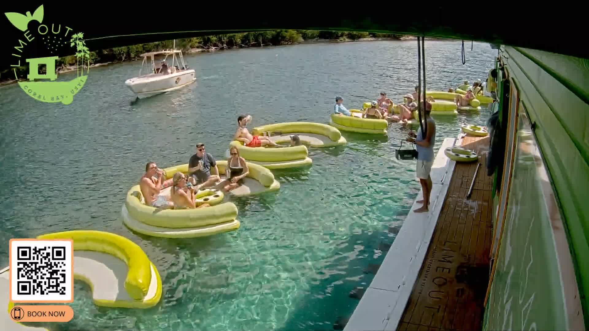 Under sunny skies, numerous people relax on yellow inflatable rafts in clear, calm turquoise water beside a large green boat, with another smaller boat visible in the distance near a tree-lined shore.