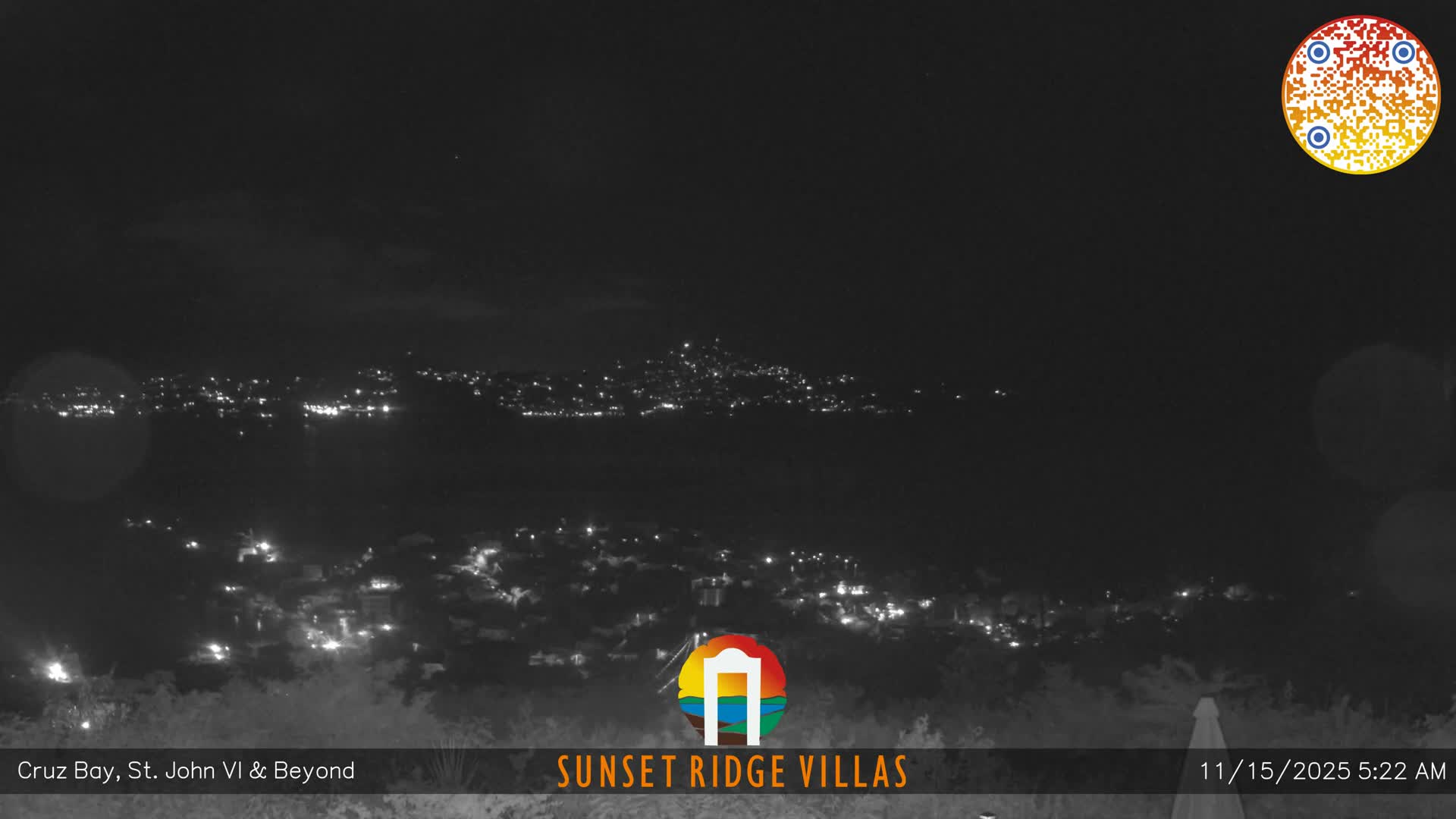 The image captures a panoramic view of a brightly lit town or city nestled along a coastline at night, under a clear or mostly clear sky.