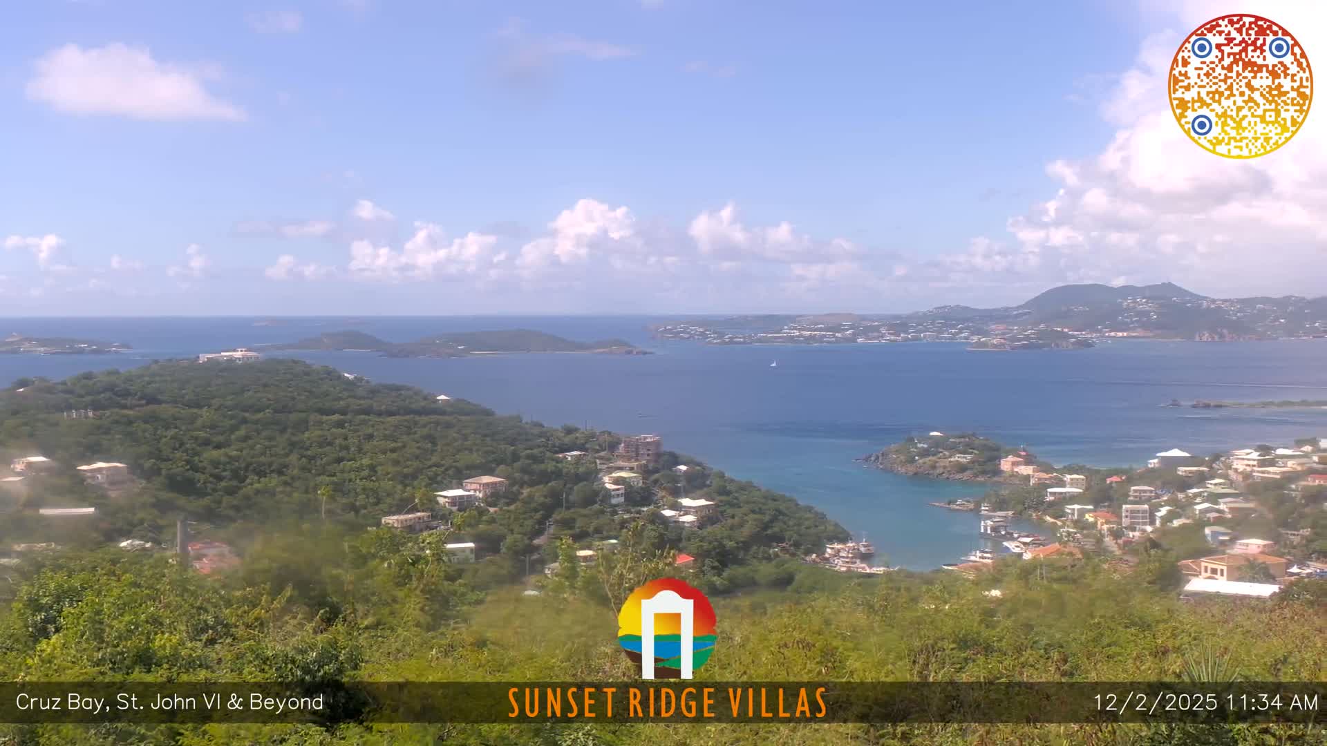 The image displays a bright, sunny day with scattered white clouds over a panoramic view of lush green tropical hills dotted with villas, overlooking a calm blue bay with boats and distant islands.