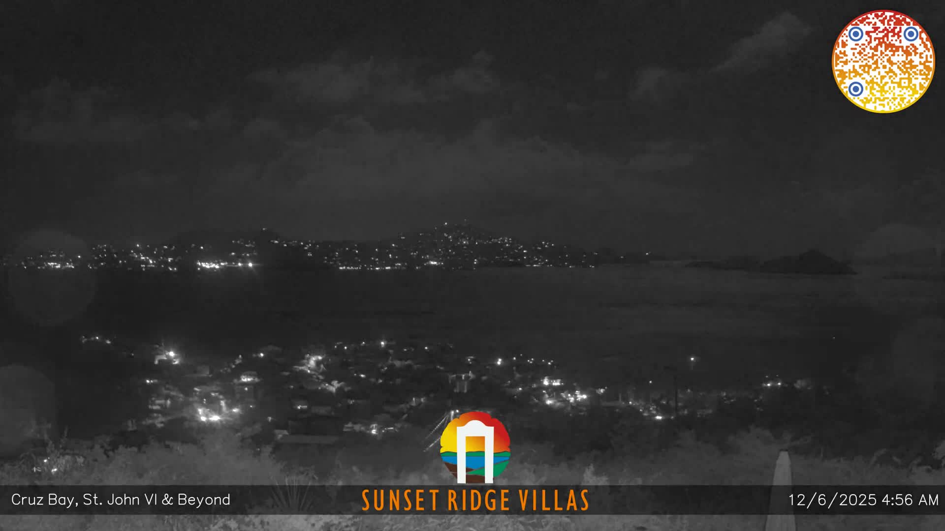 A sweeping nighttime vista reveals a densely lit coastal town sprawling across hillsides that descend to a dark bay, with distant landmasses faintly visible under an overcast sky.