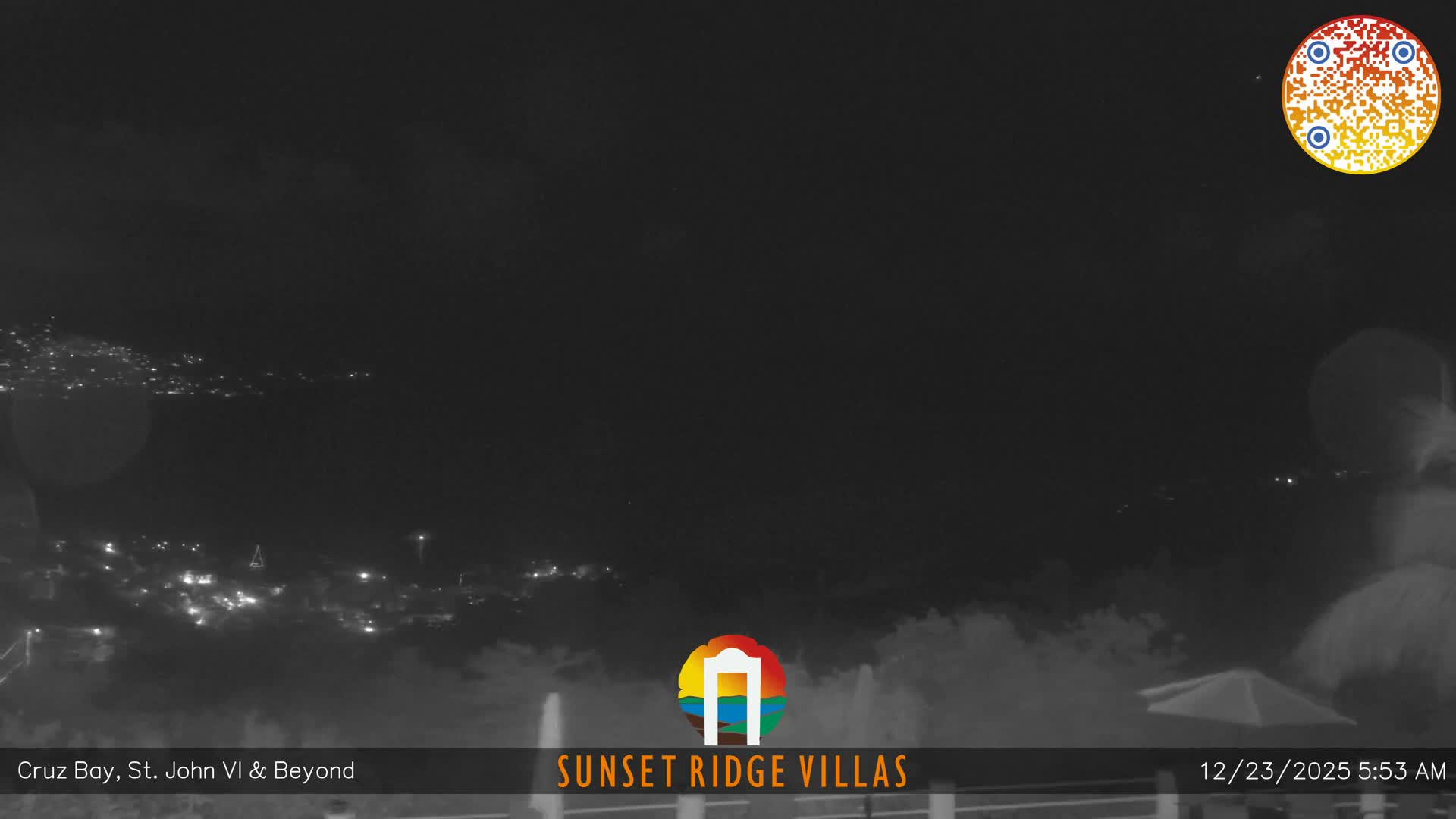A wide, dark early morning view reveals distant twinkling city lights along a dark coastline under a clear sky, with faint mist or low clouds obscuring the immediate foreground.
