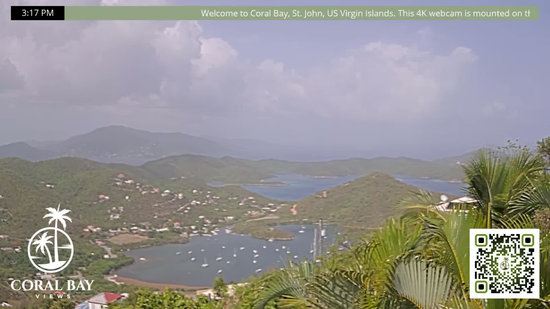 A partly cloudy day reveals a view of a calm bay filled with sailboats, nestled between verdant hills under a mostly cloudy sky.