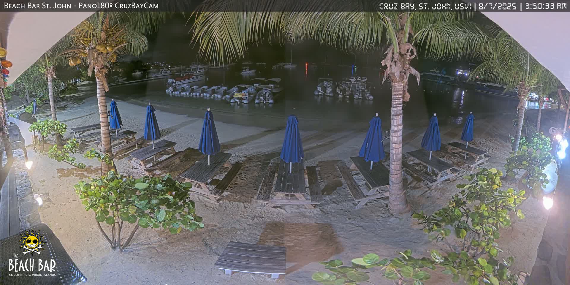 At night, several empty picnic tables with blue umbrellas sit on a sandy beach next to calm water where boats are moored.