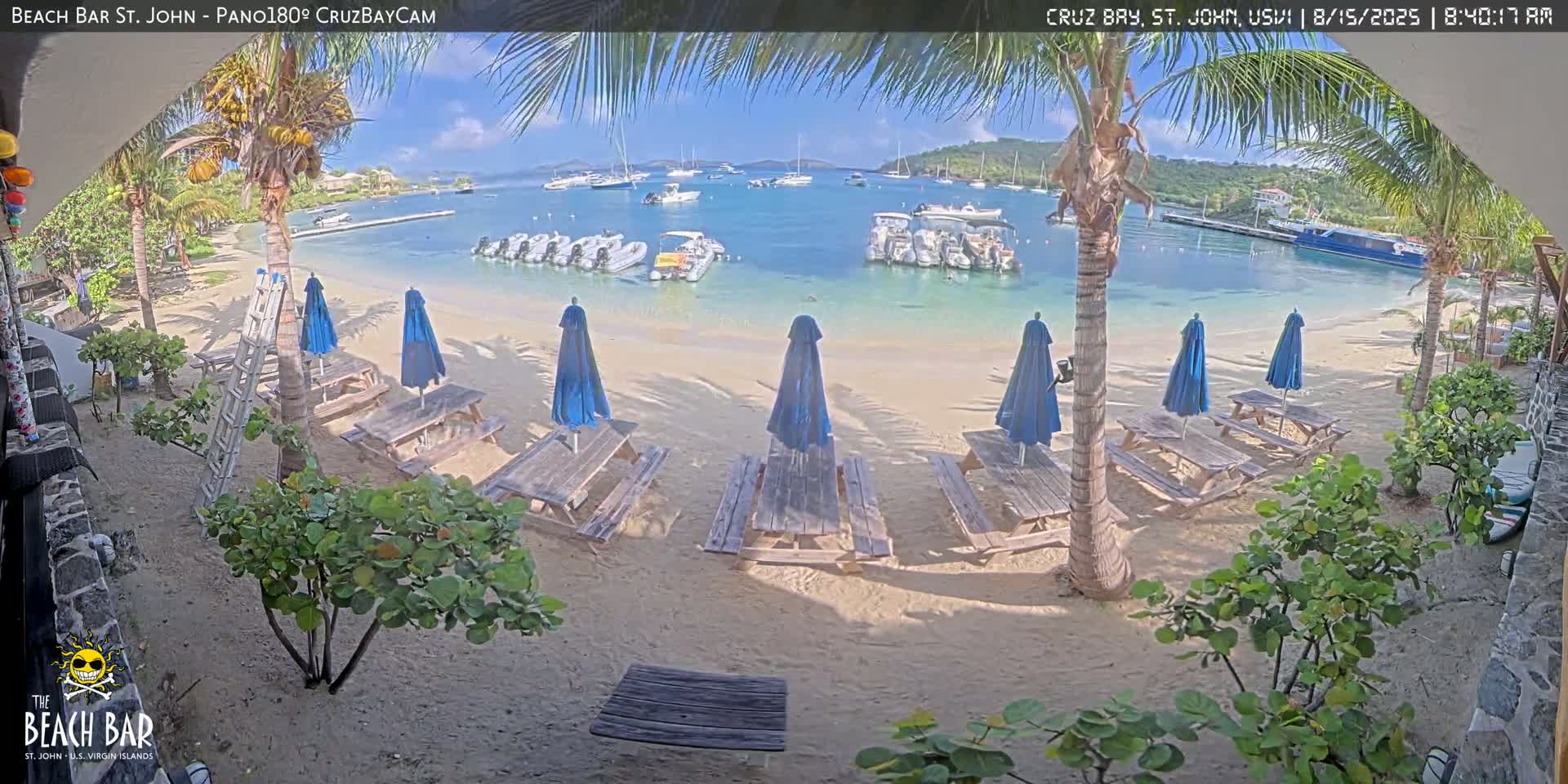 A beachside bar with picnic tables under blue umbrellas overlooks a calm bay filled with boats on an overcast day.