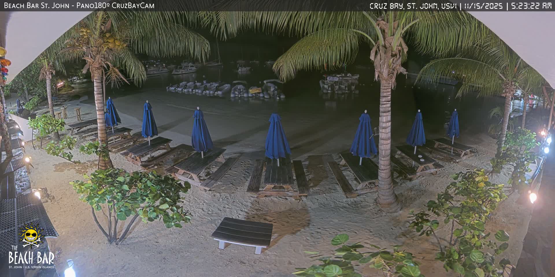 The image captures a tranquil beach at night, featuring palm trees, numerous picnic tables with closed blue umbrellas on the sand, and several small boats moored in calm water under clear skies.